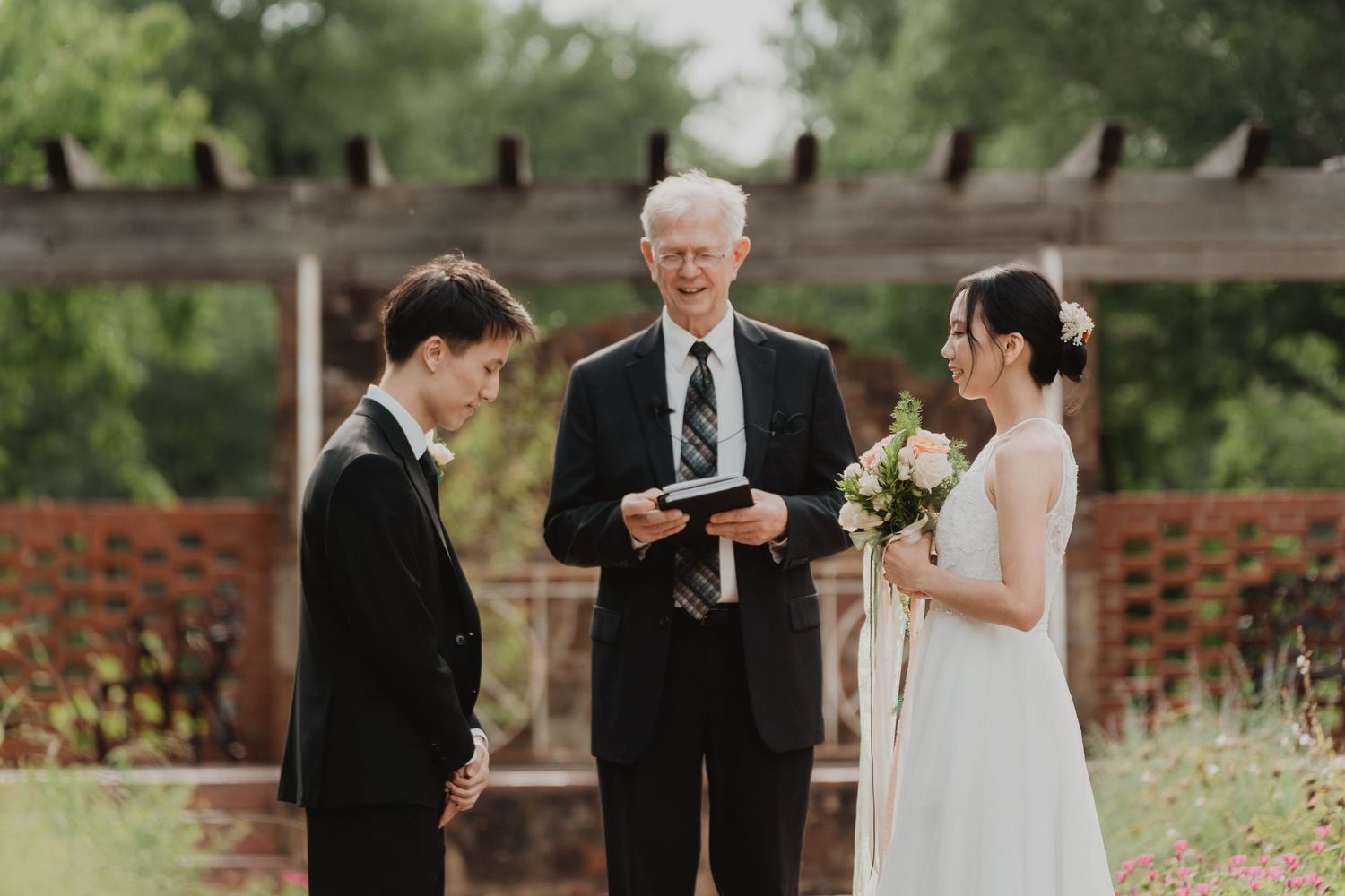 An outdoor wedding ceremony under a wooden pergola with two individuals in formal attire and an officiant reading from a book.