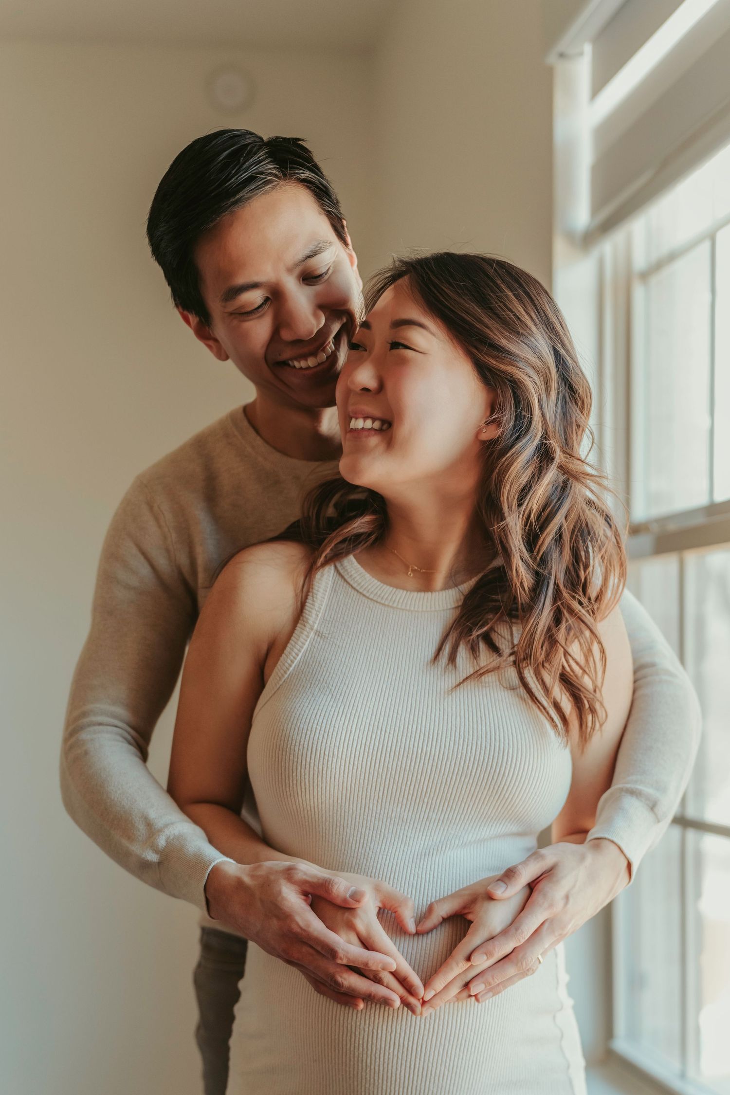 Joyful couple in casual attire embraces near a window as they form a heart shape with their hands.