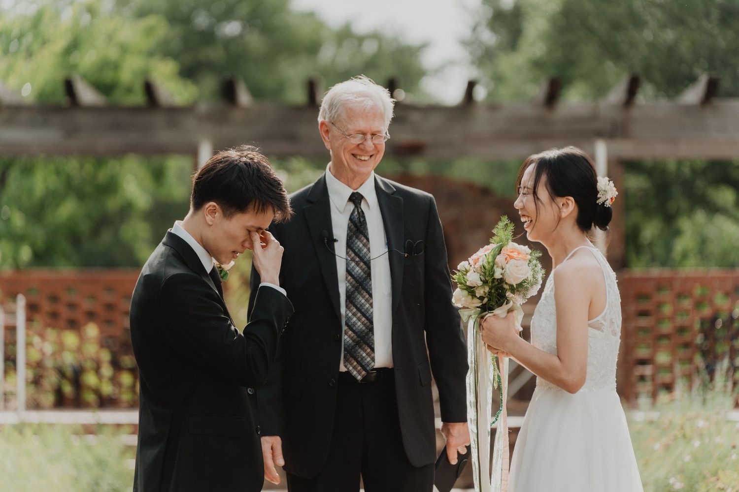 Wedding party shares joyful moments during outdoor celebration with natural greenery and brick wall backdrop.