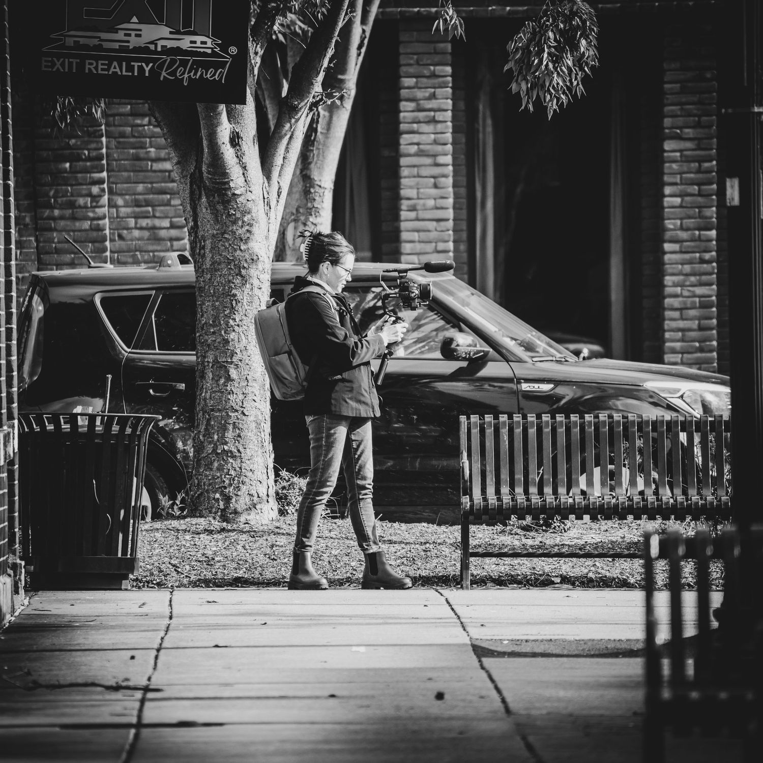 A black and white street scene showing videographer at work around downtown brick building as traffic passes by and trees stand overhead.
