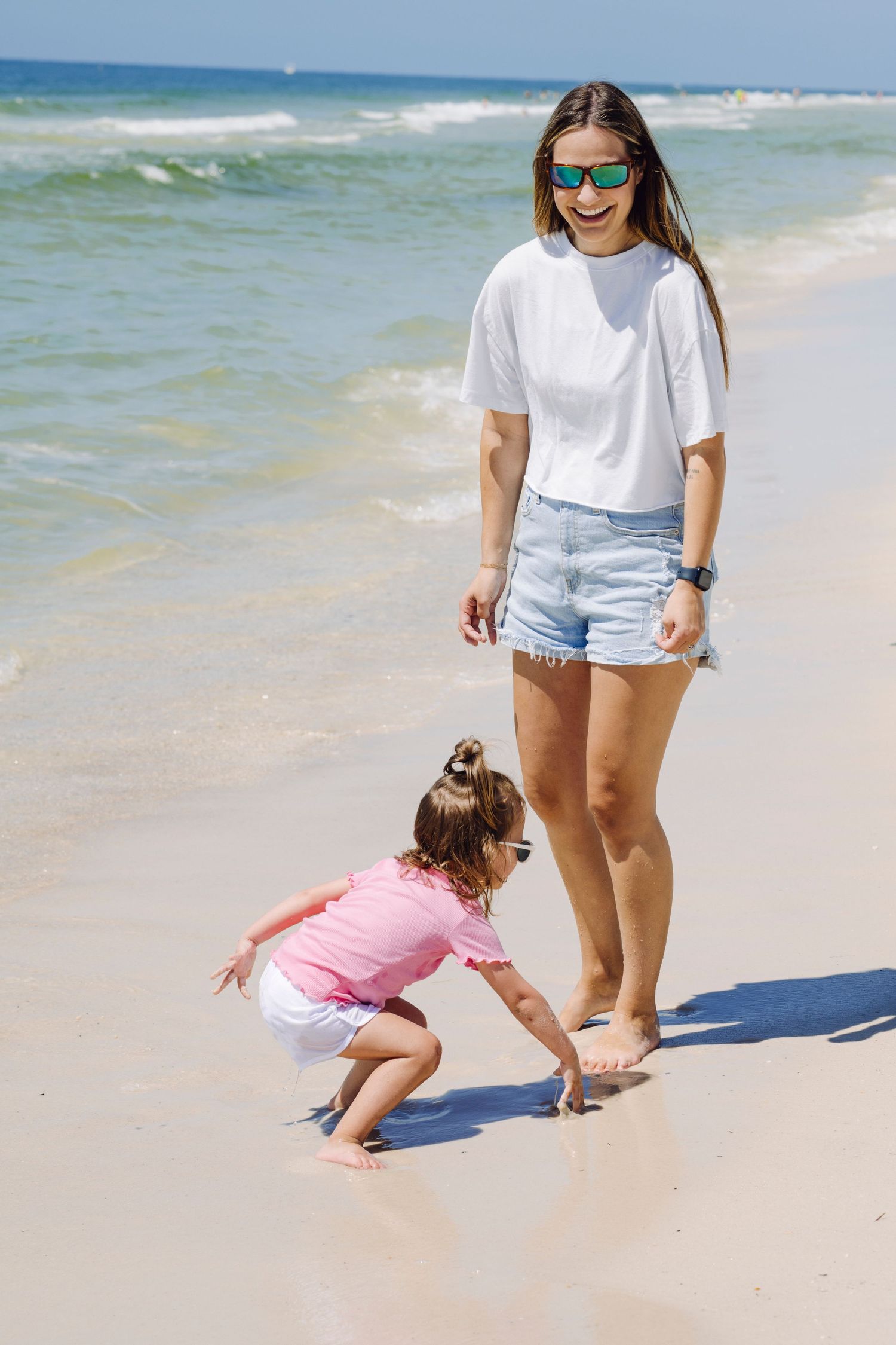 Two figures in casual summer attire explore a sandy shoreline where gentle waves meet pristine white beach.
