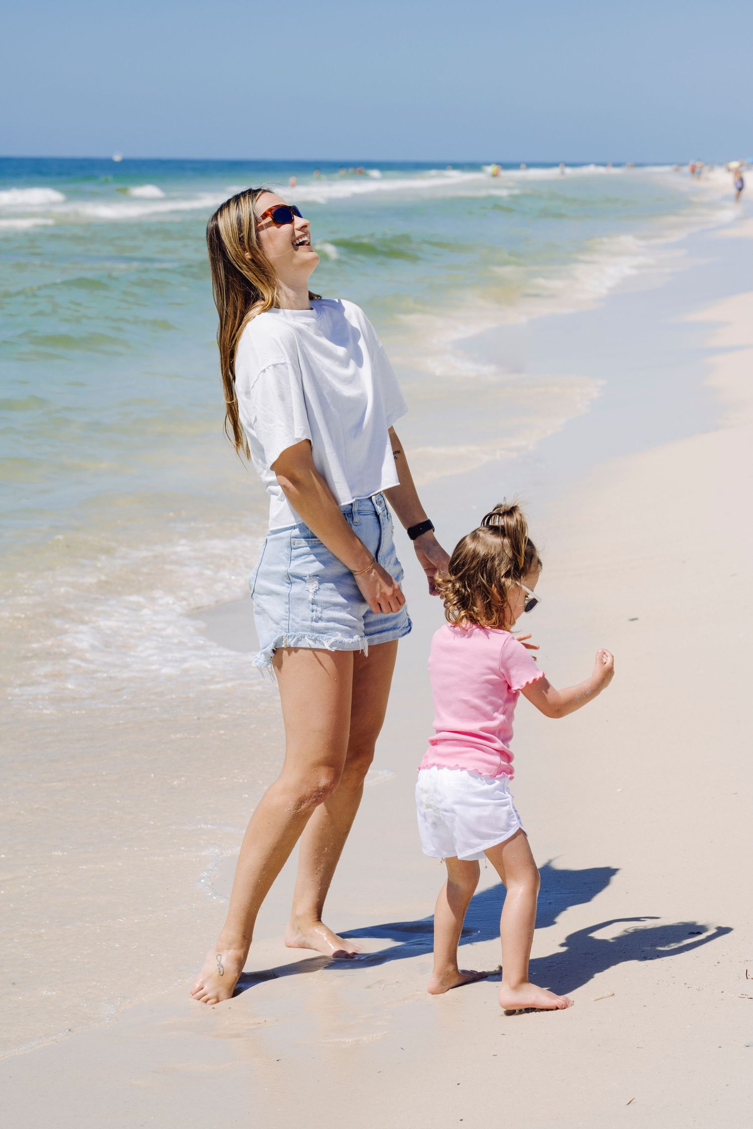 Two figures enjoy a sunny day walking barefoot along a white sandy beach with turquoise ocean waves in the background.