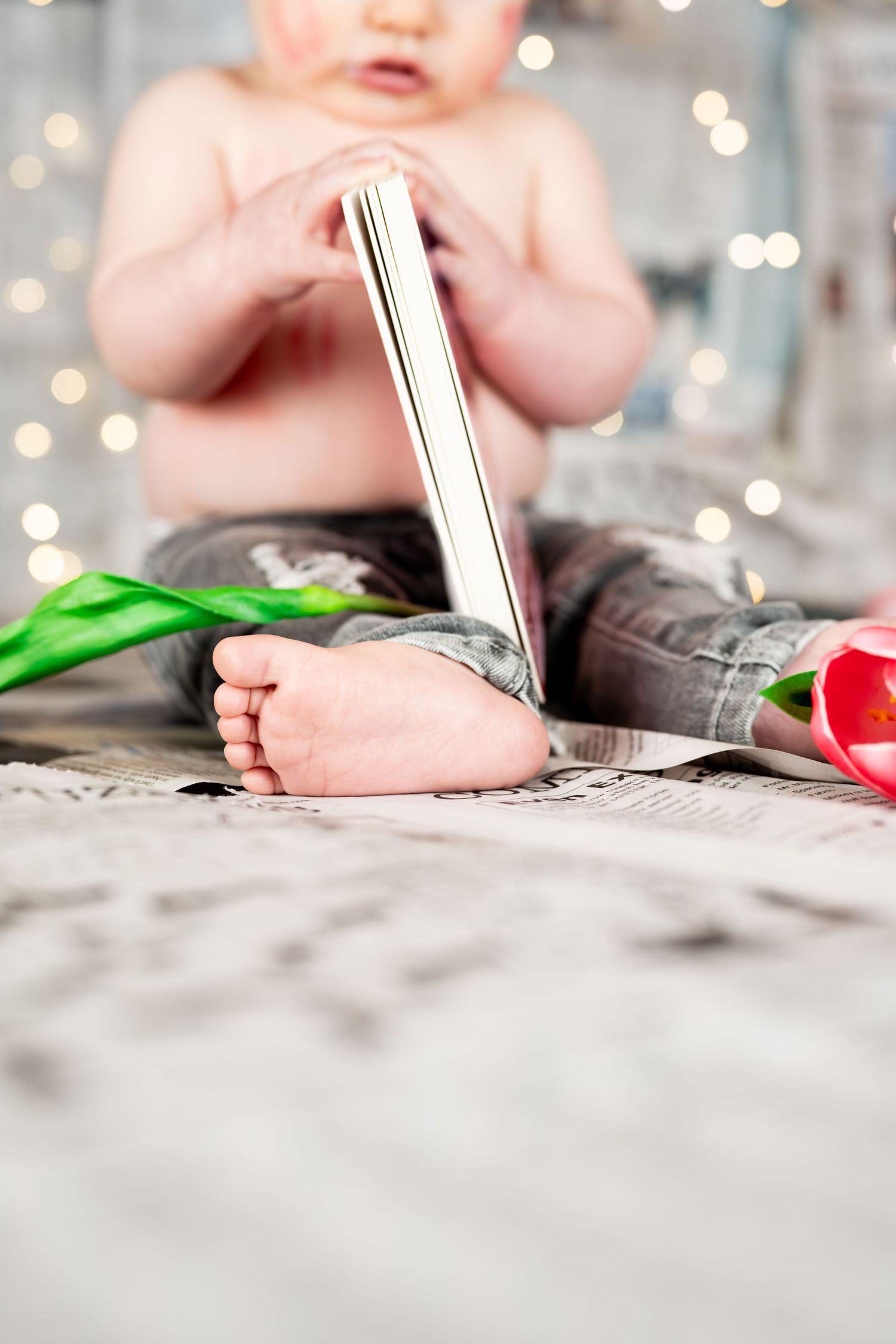 Baby in jeans sitting with a book and pink tulip on newspaper, with bokeh lights in the background.