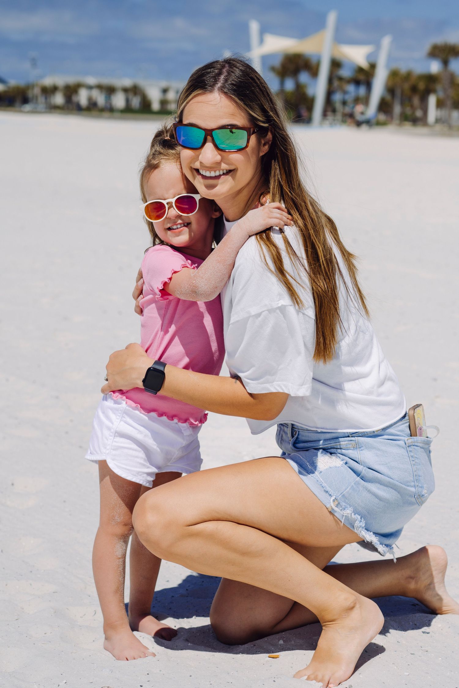 Two people wearing sunglasses embrace and smile on a sunny beach, dressed in summer attire.