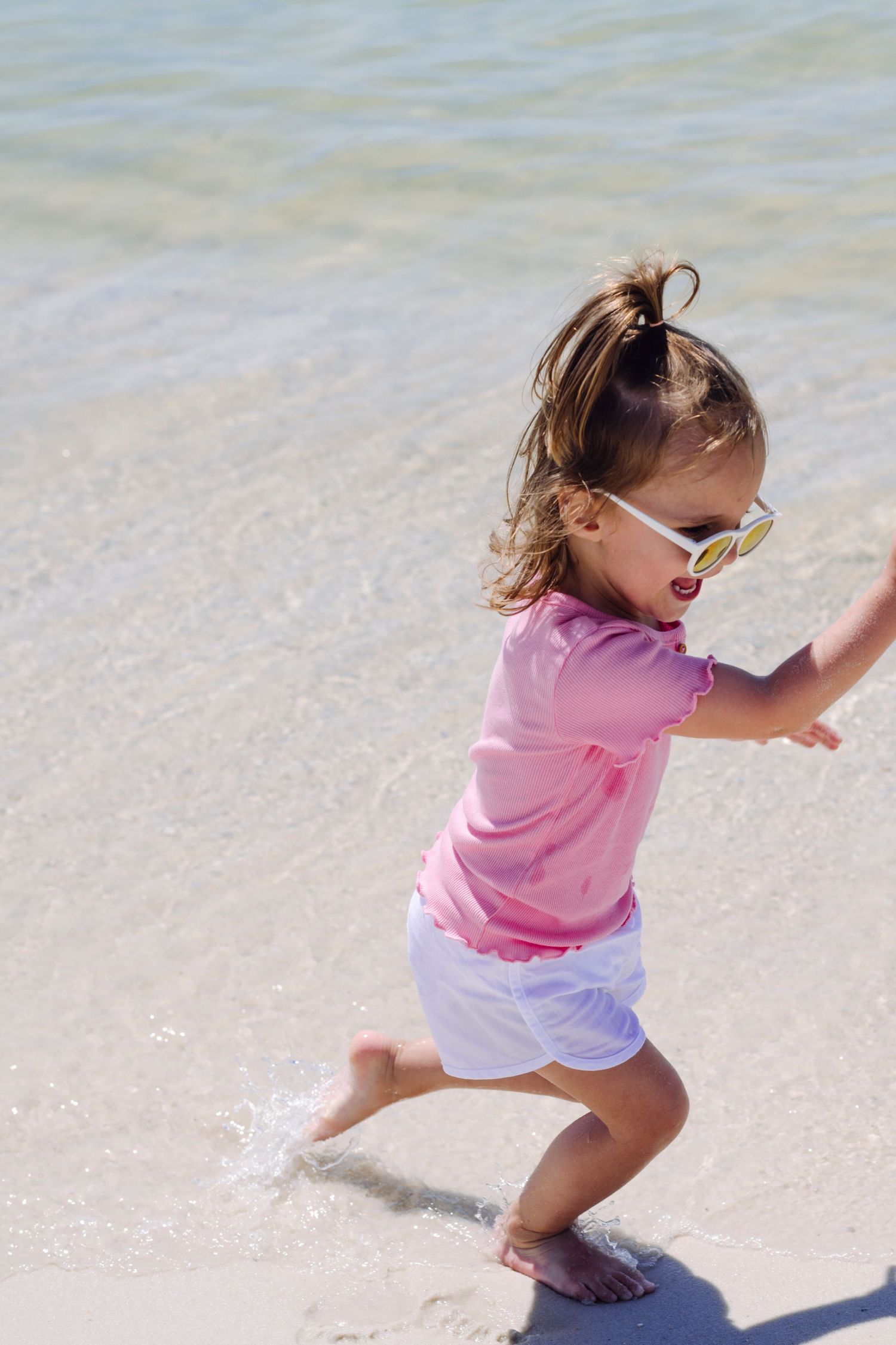 Small child in pink shirt and white shorts runs playfully along a sandy beach on a bright sunny day.