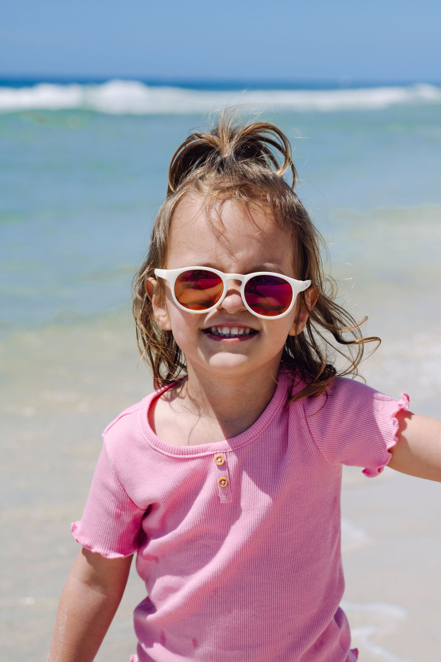 Child wearing pink shirt and sunglasses plays joyfully at a sunny beach with ocean waves in background.