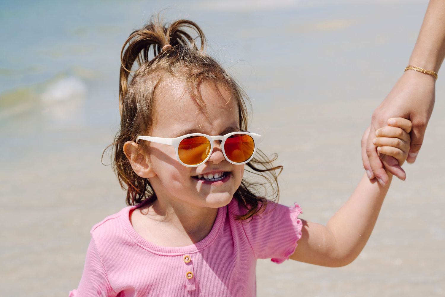 Happy child in pink shirt and sunglasses playing at the beach while holding someone's hand on a sunny day.