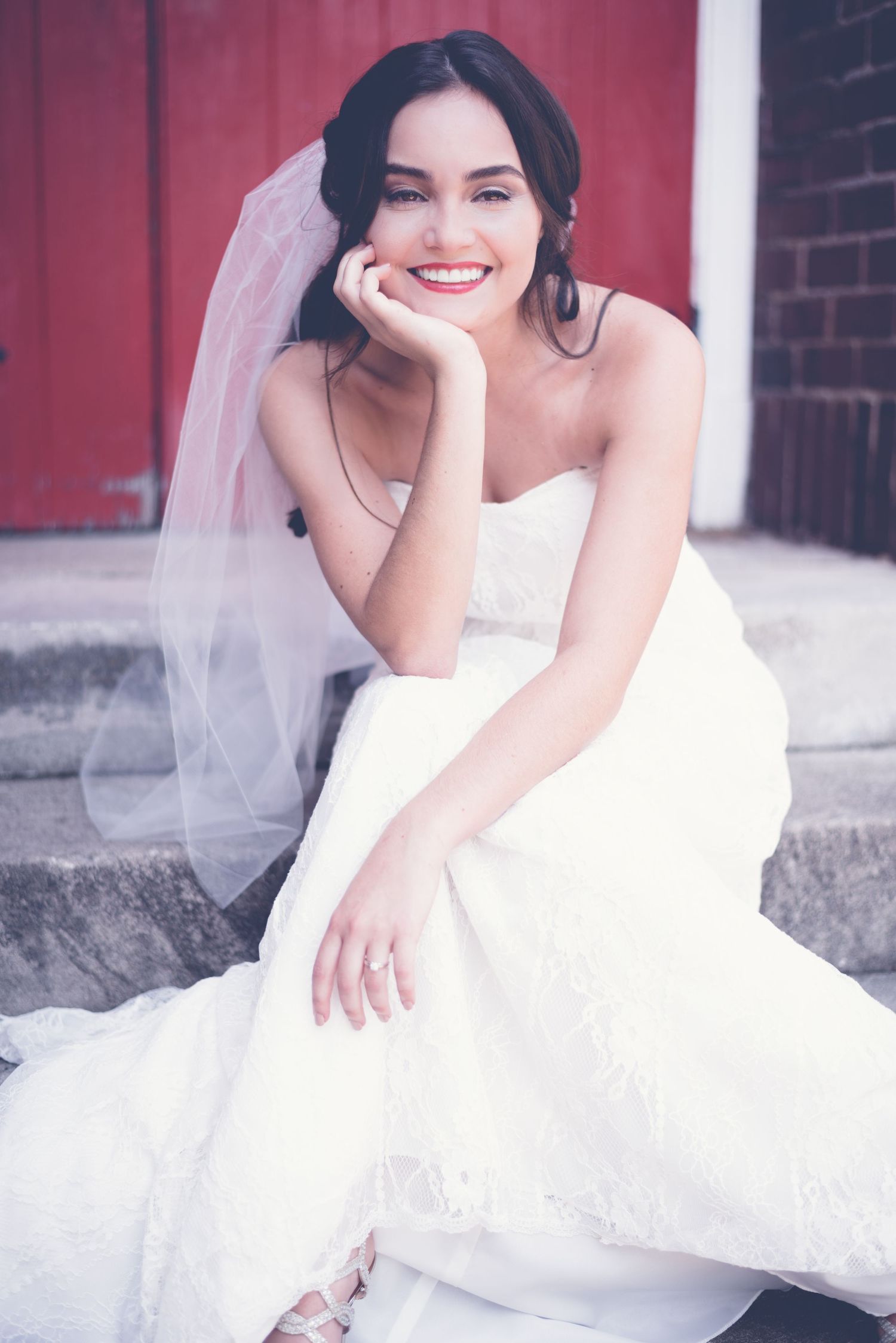 A bride in a classic white wedding gown and veil sits casually on stone steps near a red church door, in vintage style.