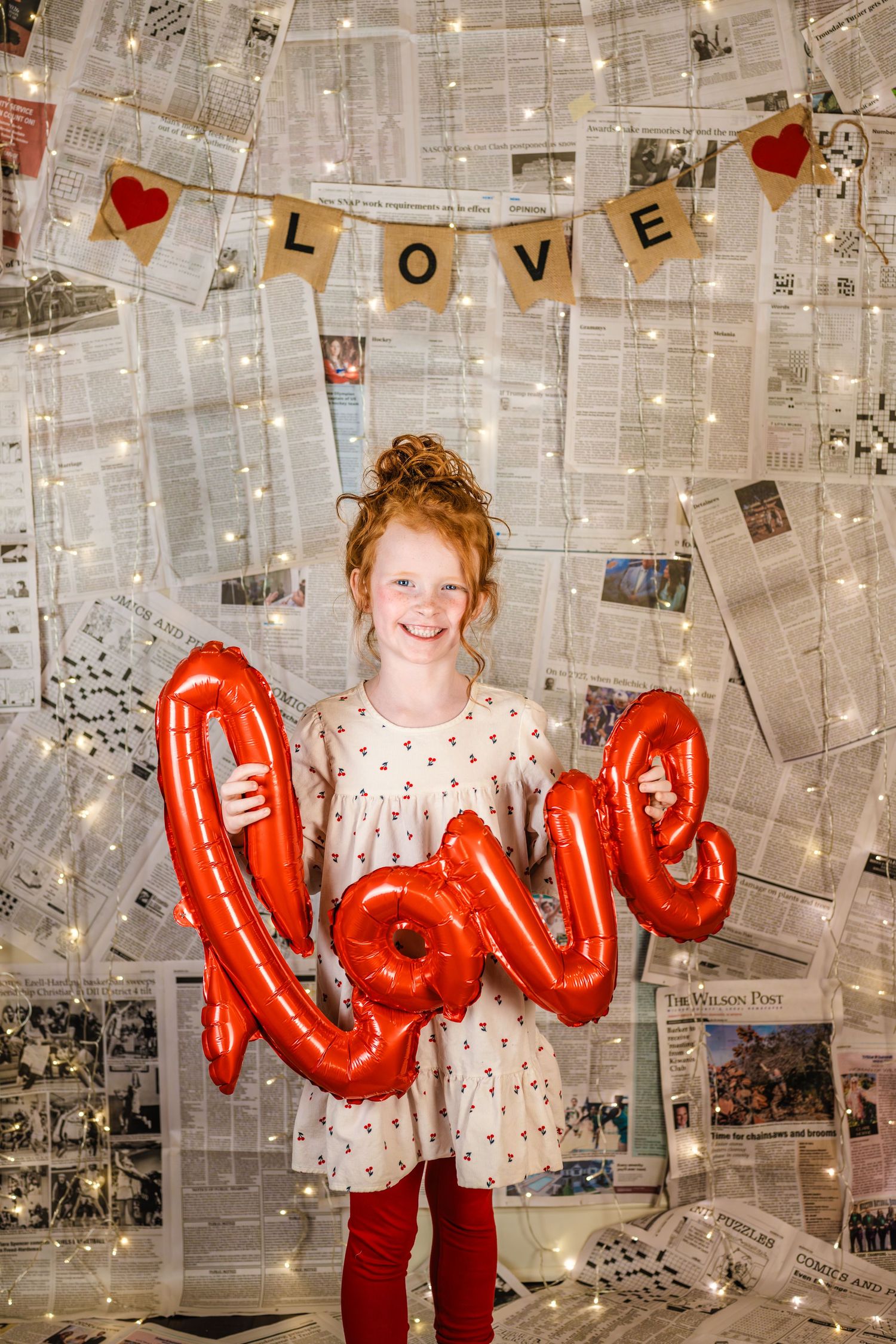 Smiling redhead girl holds red love balloon in front of newspaper wall with fairy lights and LOVE banner.