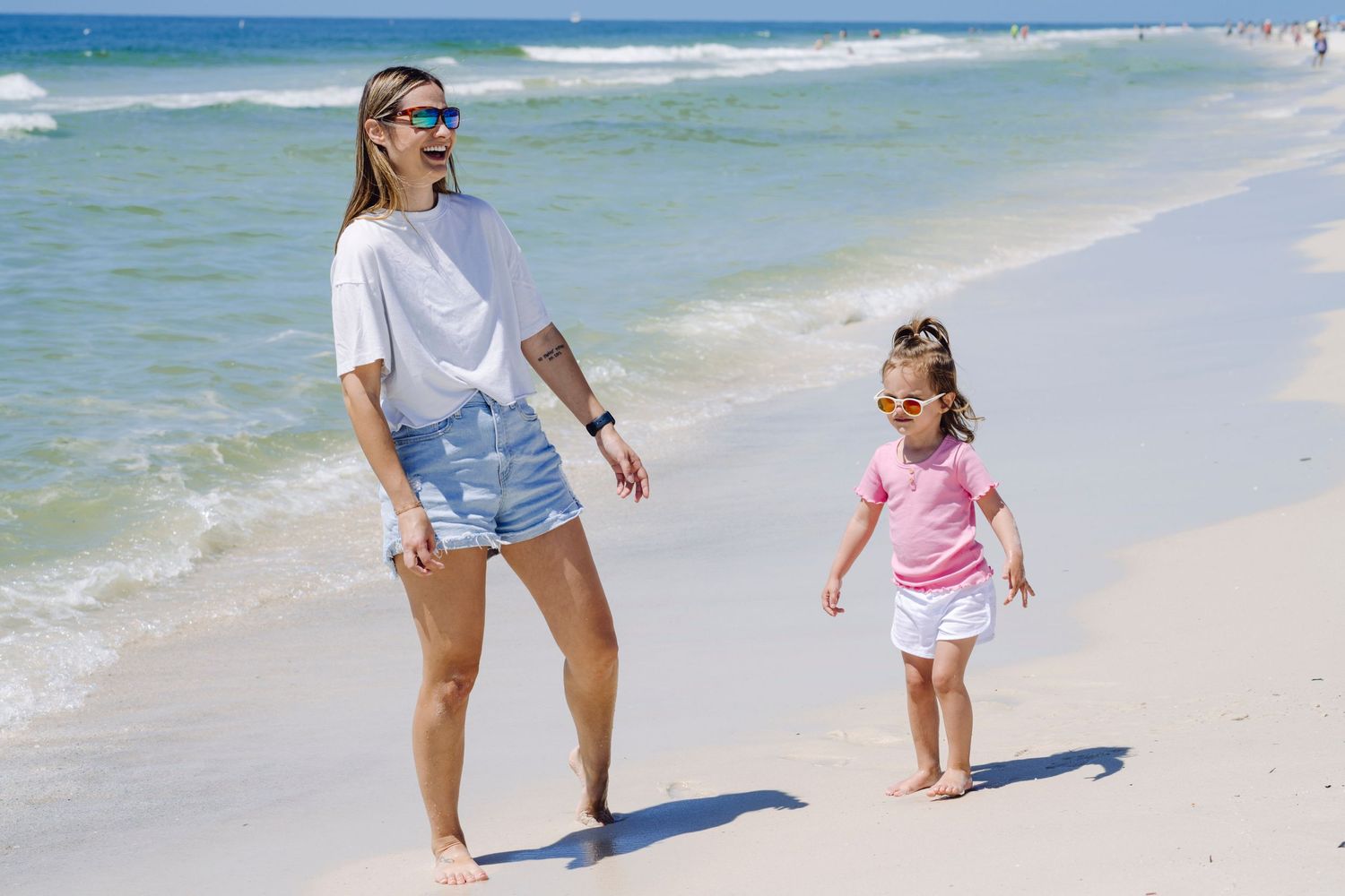 Two people walk together along a sunny beach with turquoise waves and white sand in the background.