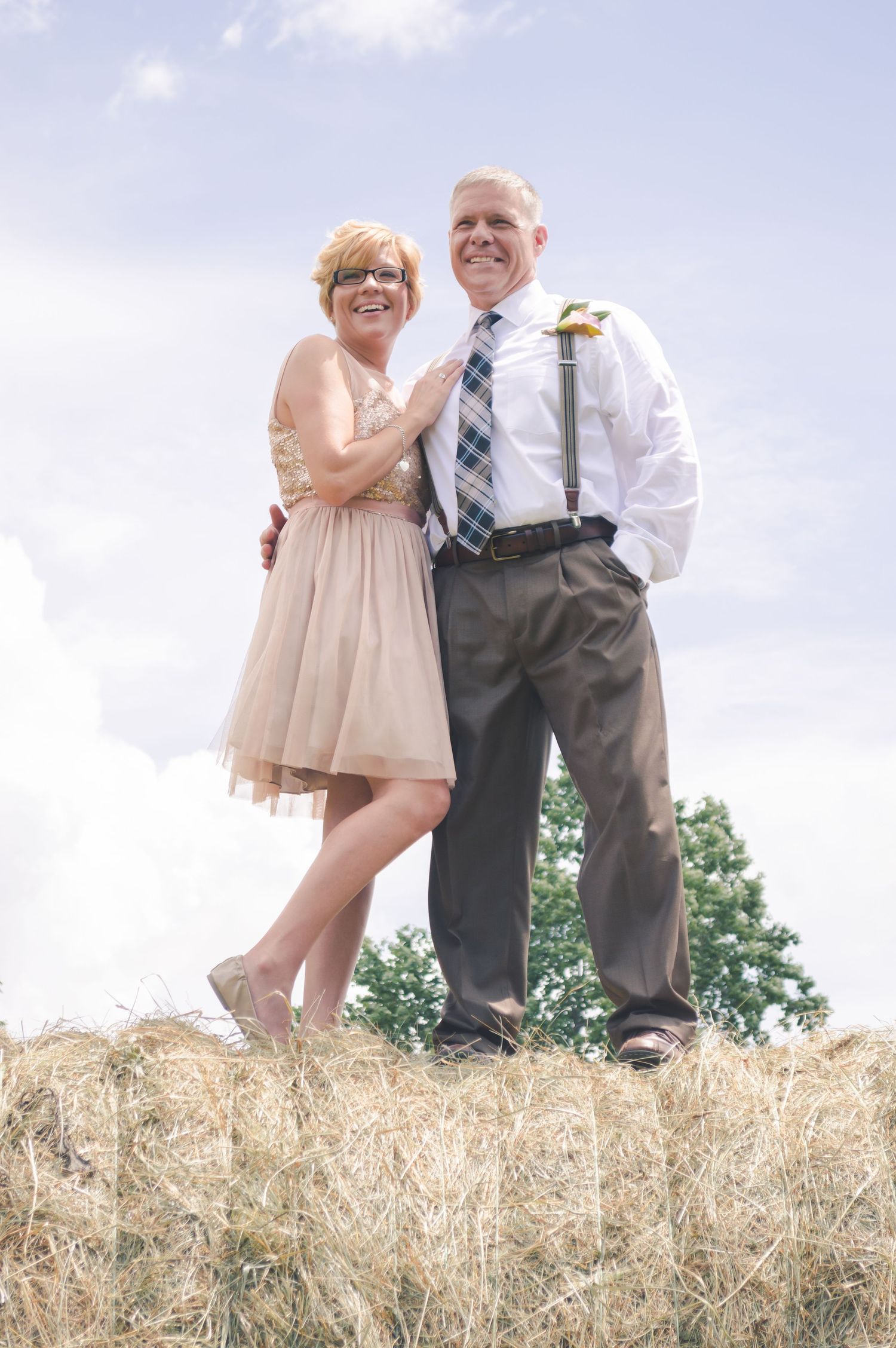 A happy couple poses together on top of a hay bale, with the woman in a blush dress and man in dress shirt and tie.