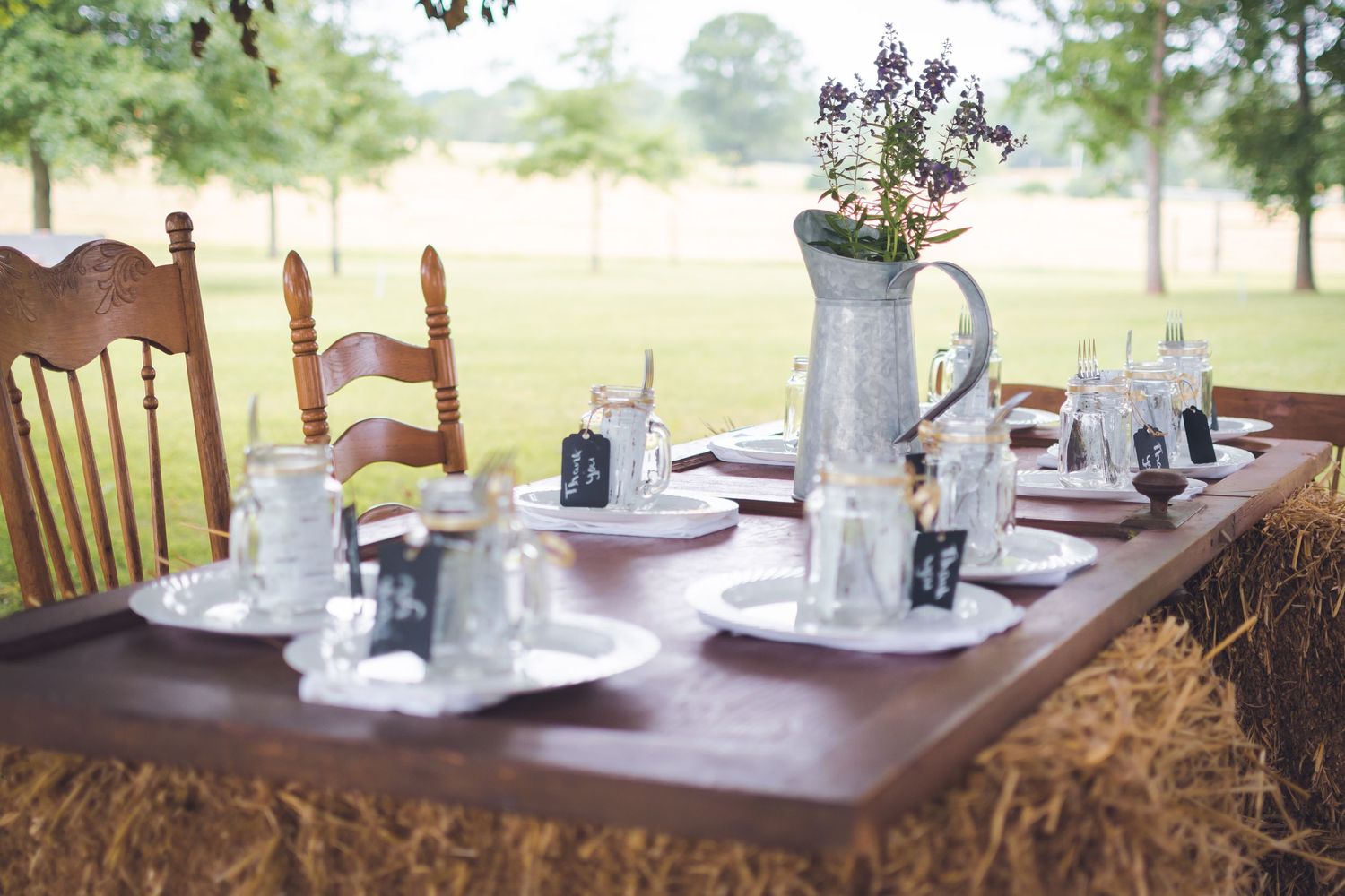 Rustic outdoor table setting with vintage white teacups and lavender flowers in a metal pitcher on wooden surface.