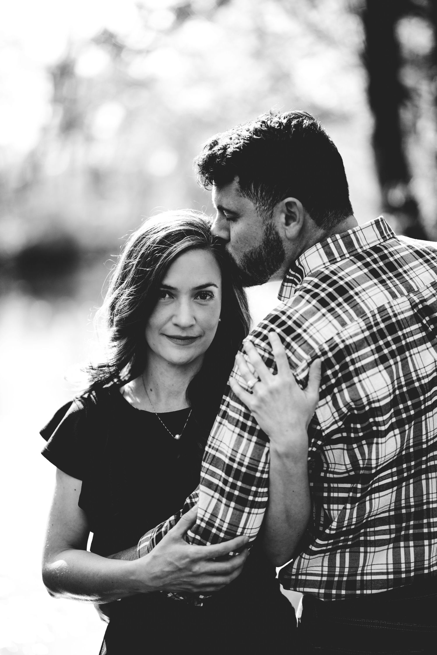 Black and white shot of a couple during a lifestyle creates a timeless look of intimate love as he kisses her on the head.