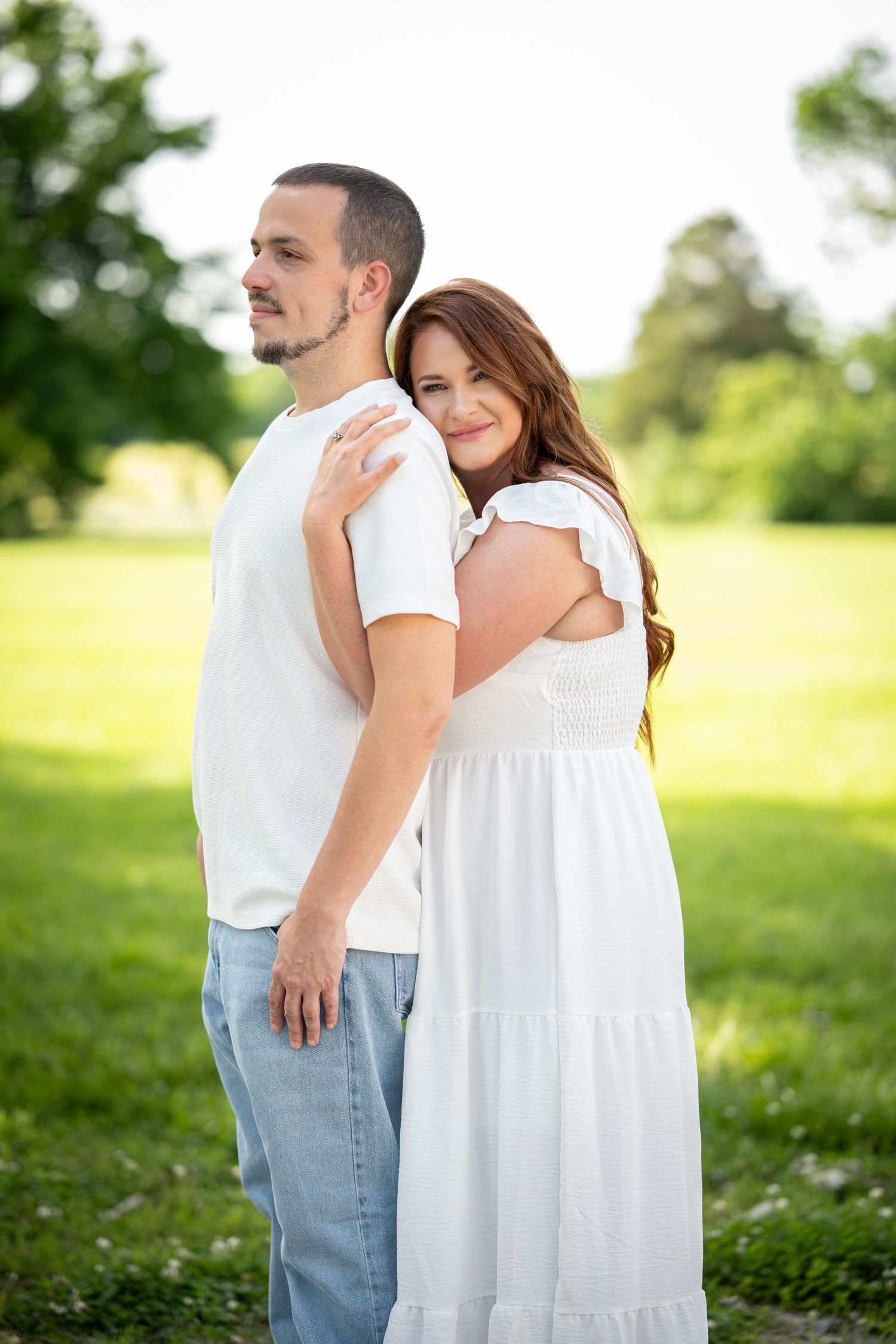 A couple in white outfits embrace while standing in a sunny green field on a summer day.