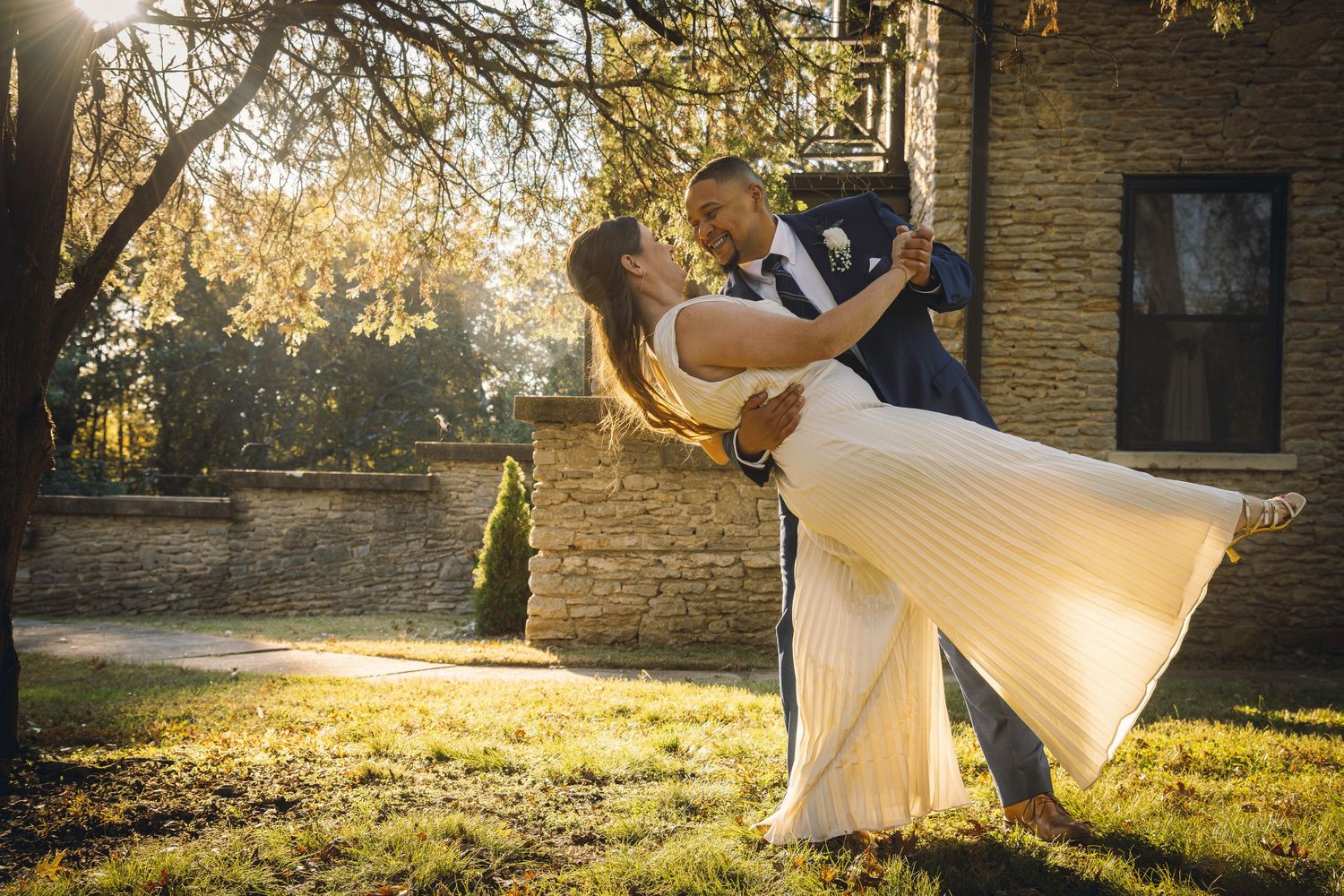 A groom dips his bride in a sunlit garden, her white dress flowing, near a stone building surrounded by autumn trees.