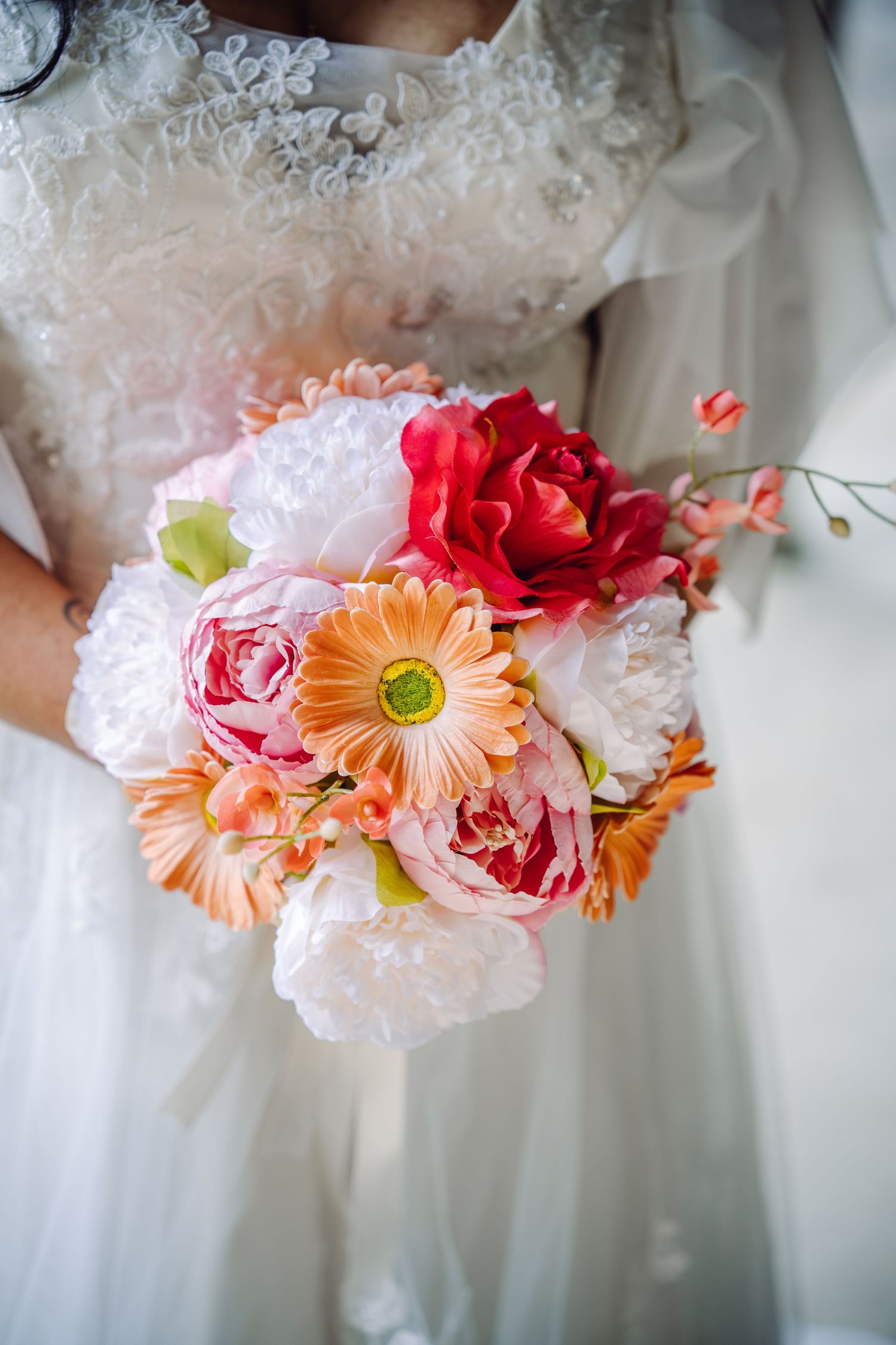 Bride in white lace gown holding a vibrant wedding bouquet with peonies, gerbera daisies, and roses.