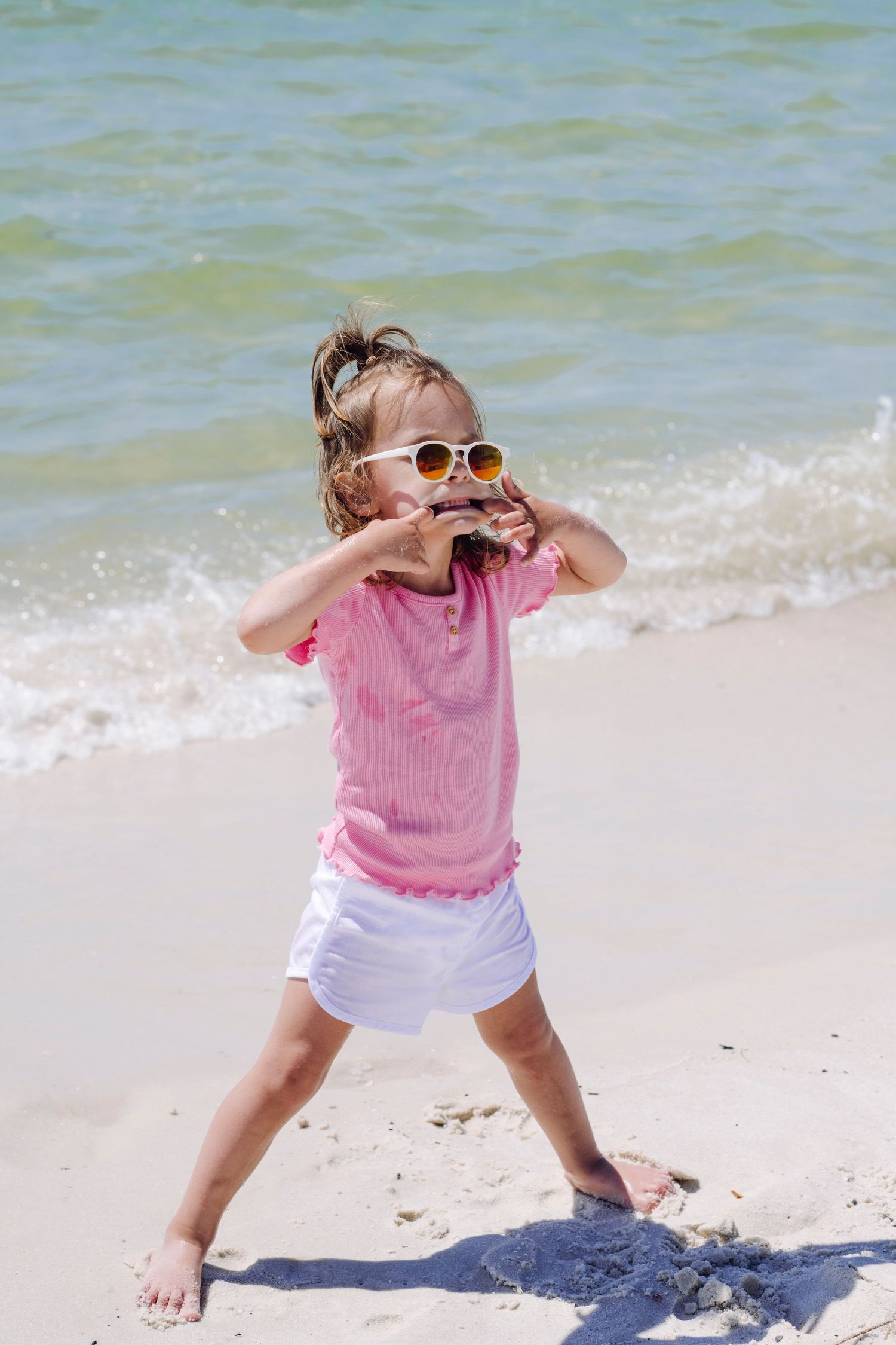 A series of photos showing a young child in a pink shirt playing and dancing energetically on a sunny beach shoreline.