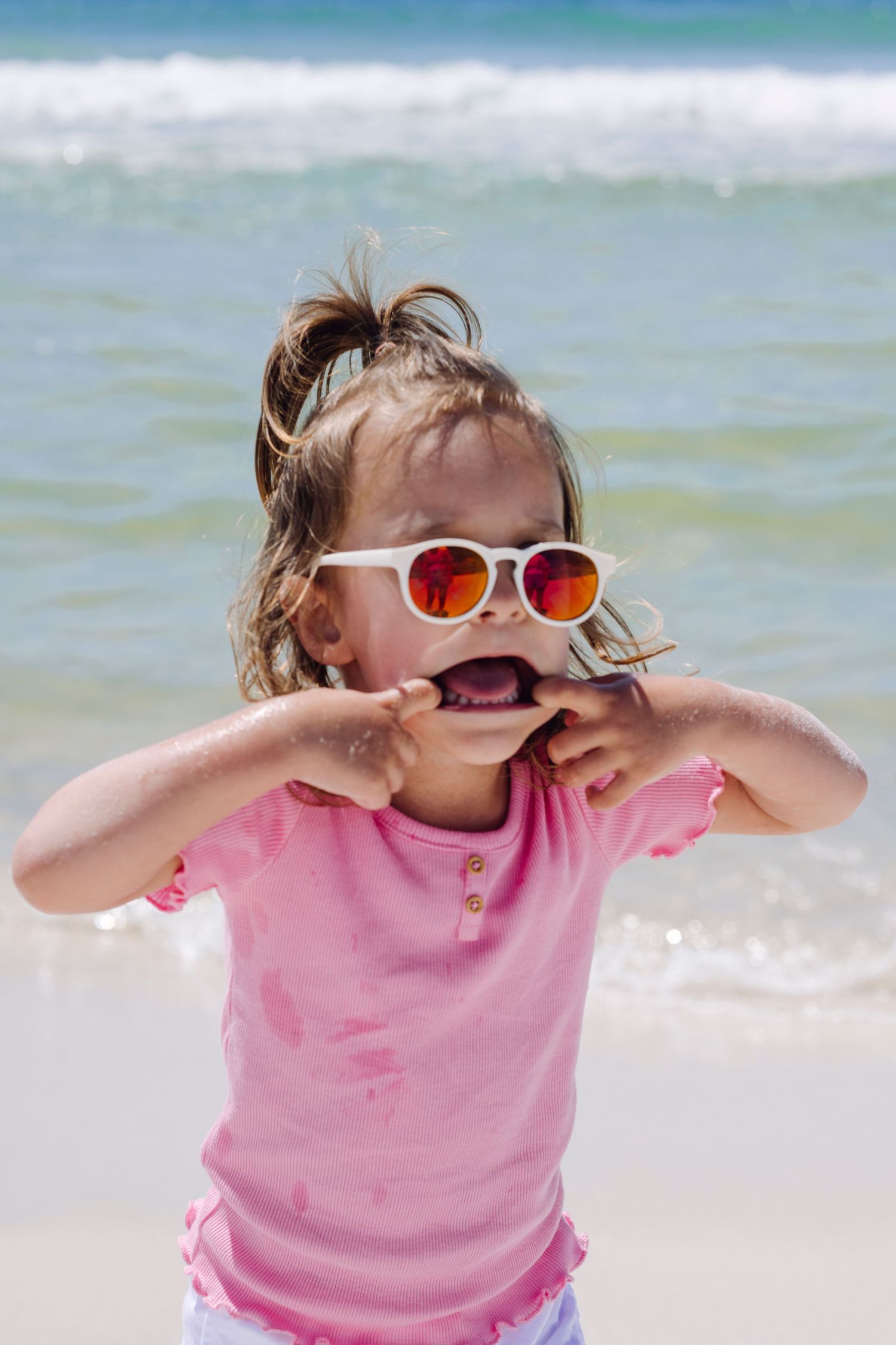 Young child wearing pink shirt and white sunglasses makes silly face while playing at sunny beach.