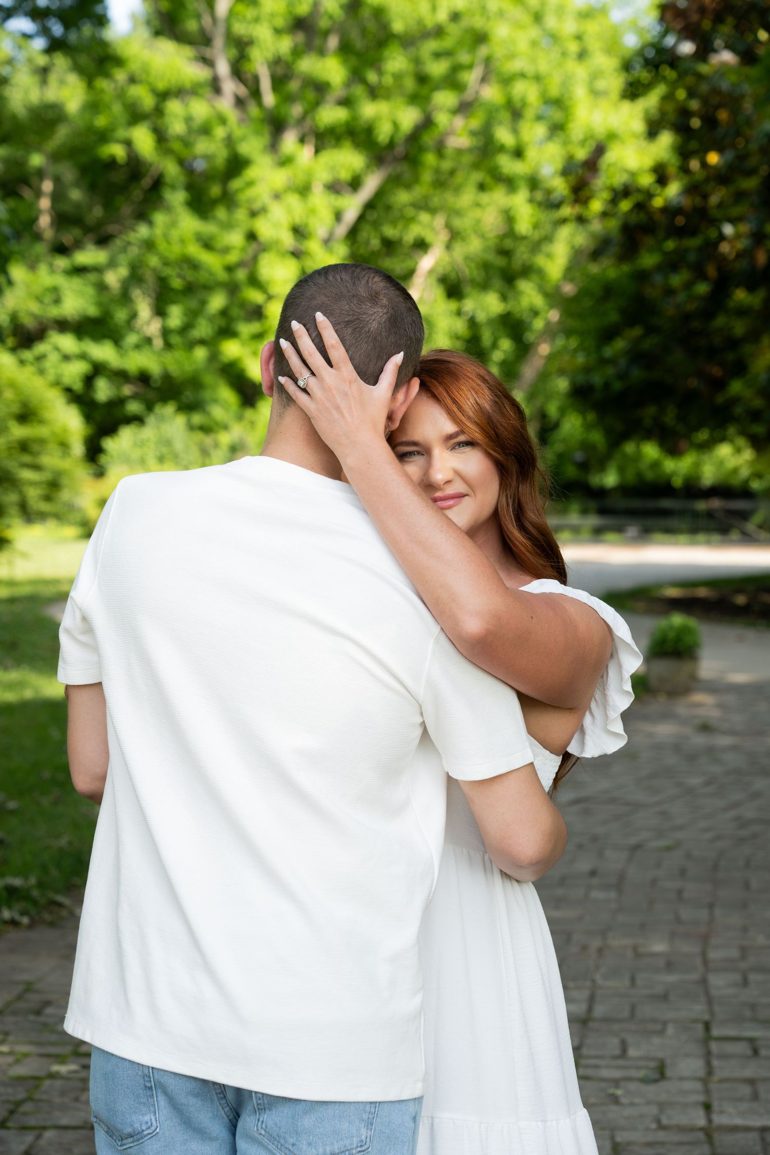 A couple shares a tender embrace on a brick pathway surrounded by lush green trees in a summer garden setting.
