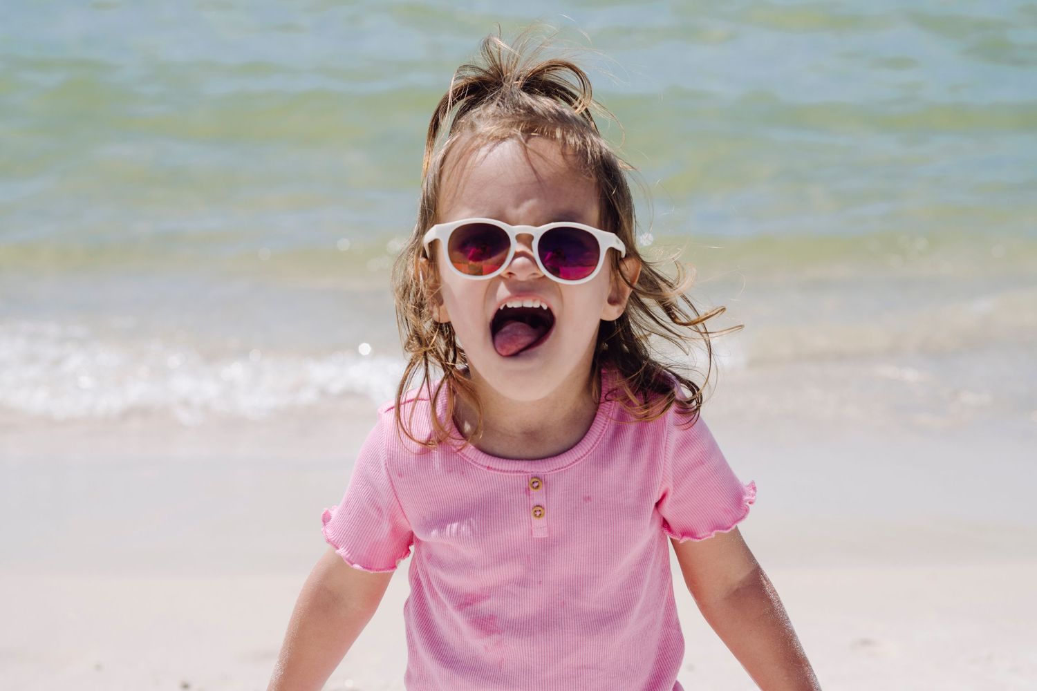 Child at beach wearing pink shirt and sunglasses appears excited with open mouth expression against ocean backdrop.