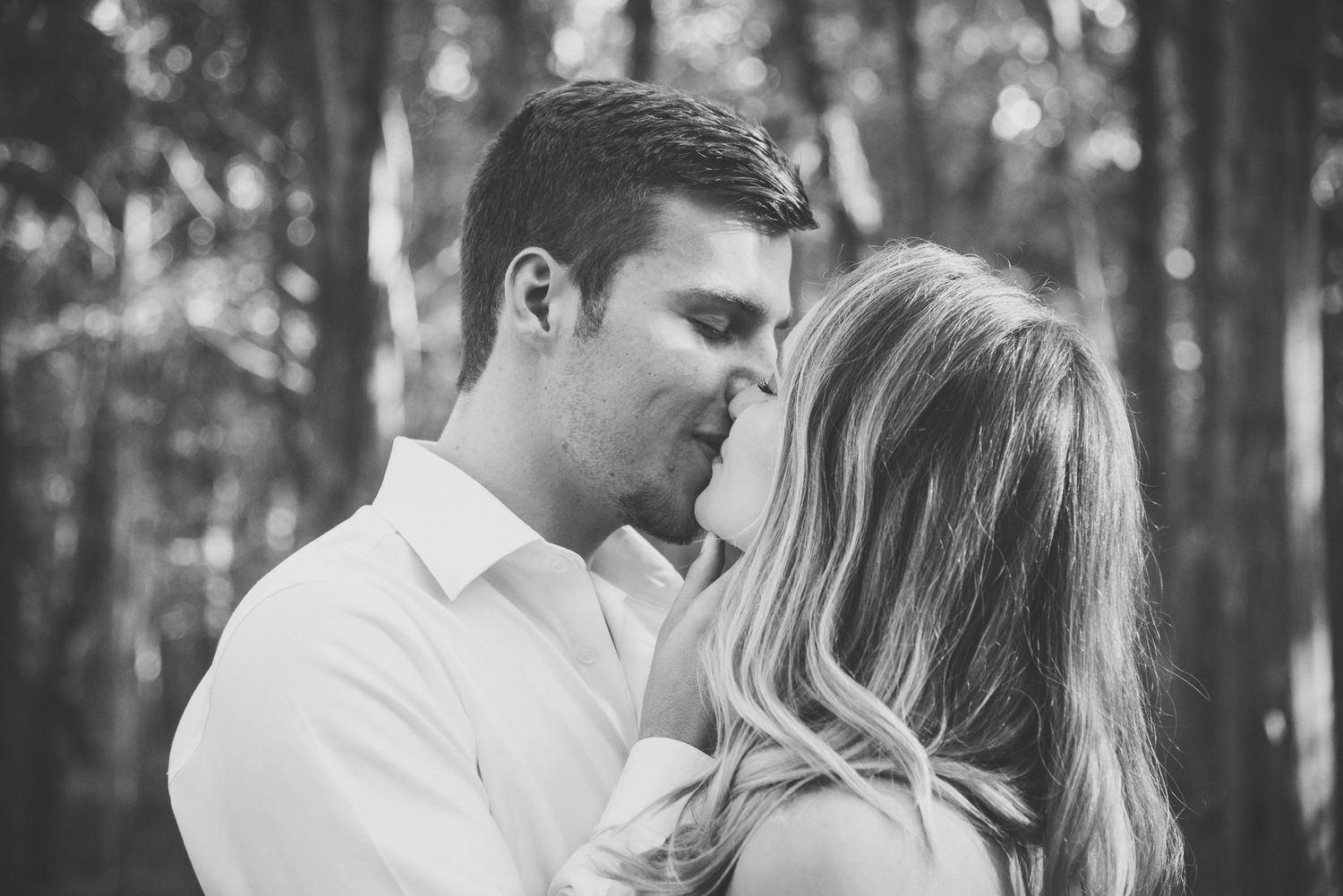 Black and white romantic photo of couple embracing in a forest, as the man gently lifts the woman's chin into to kiss her.