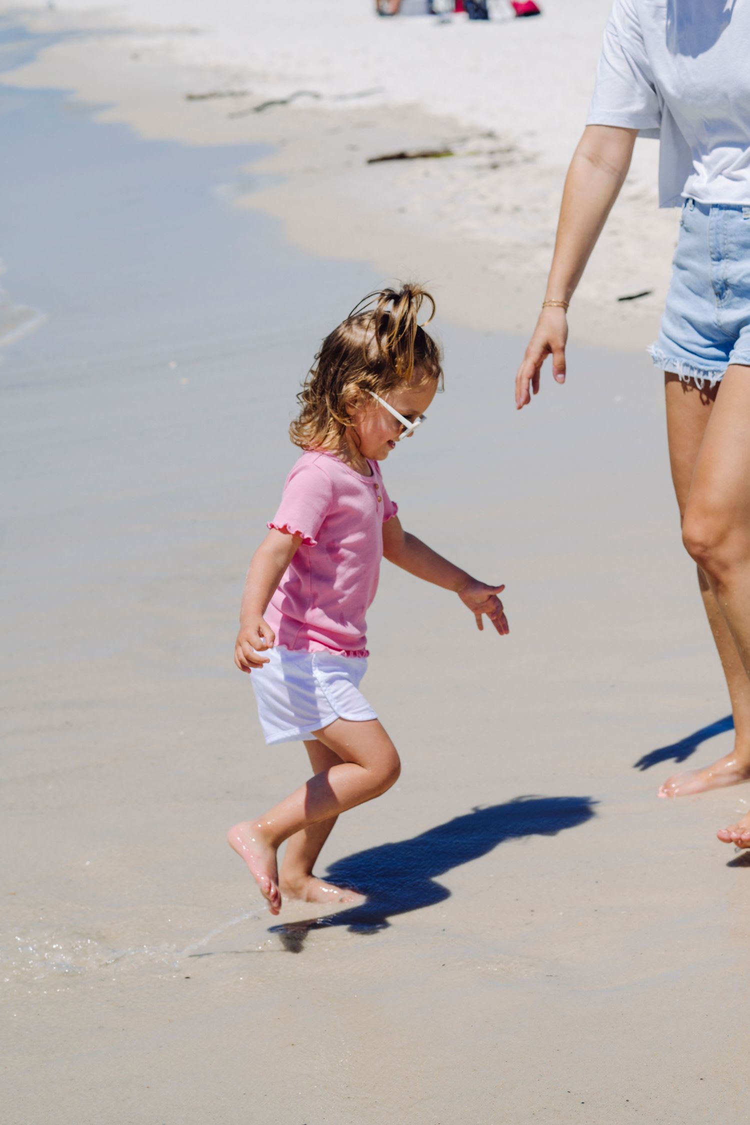 A child in a pink shirt and white shorts plays on a sandy beach while a guardian stands nearby.