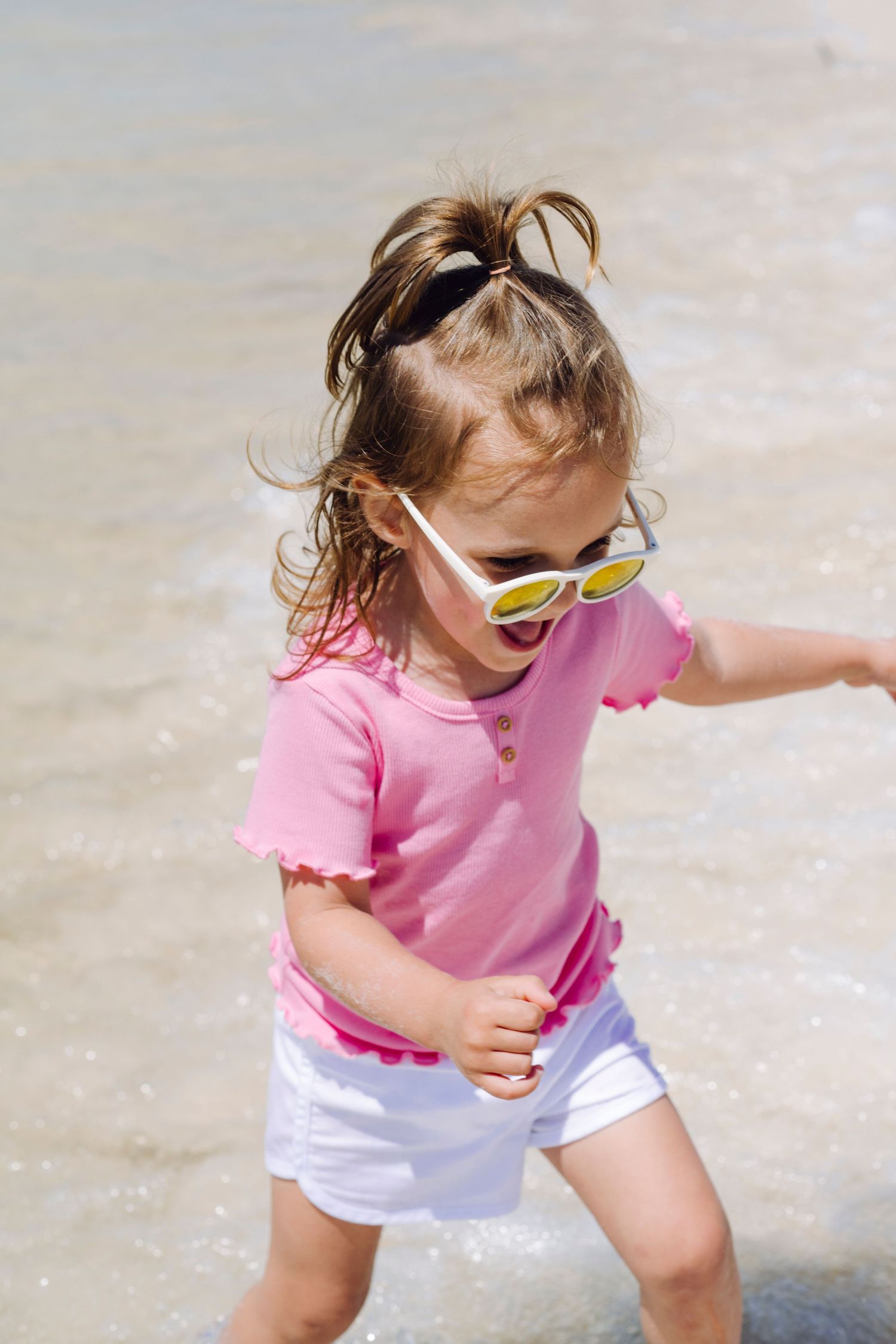 Small child in pink shirt and yellow sunglasses plays energetically outdoors on a sunny day.