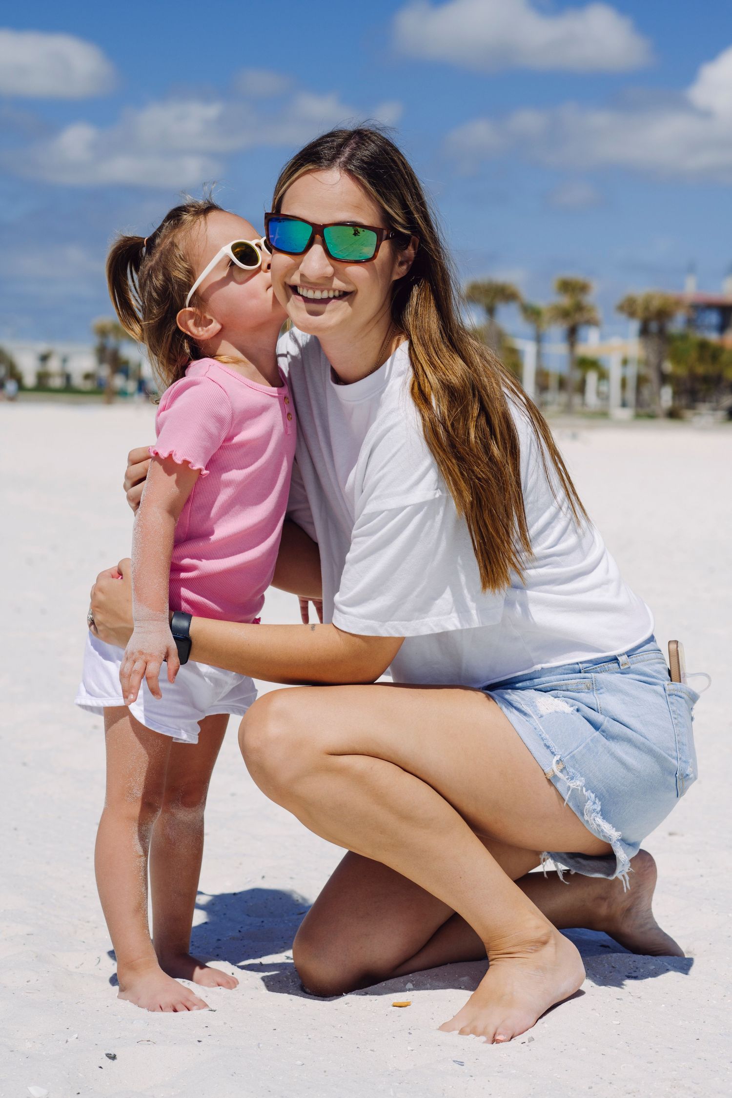 A parent and child share a sweet moment on a sunny beach while wearing colorful sunglasses.