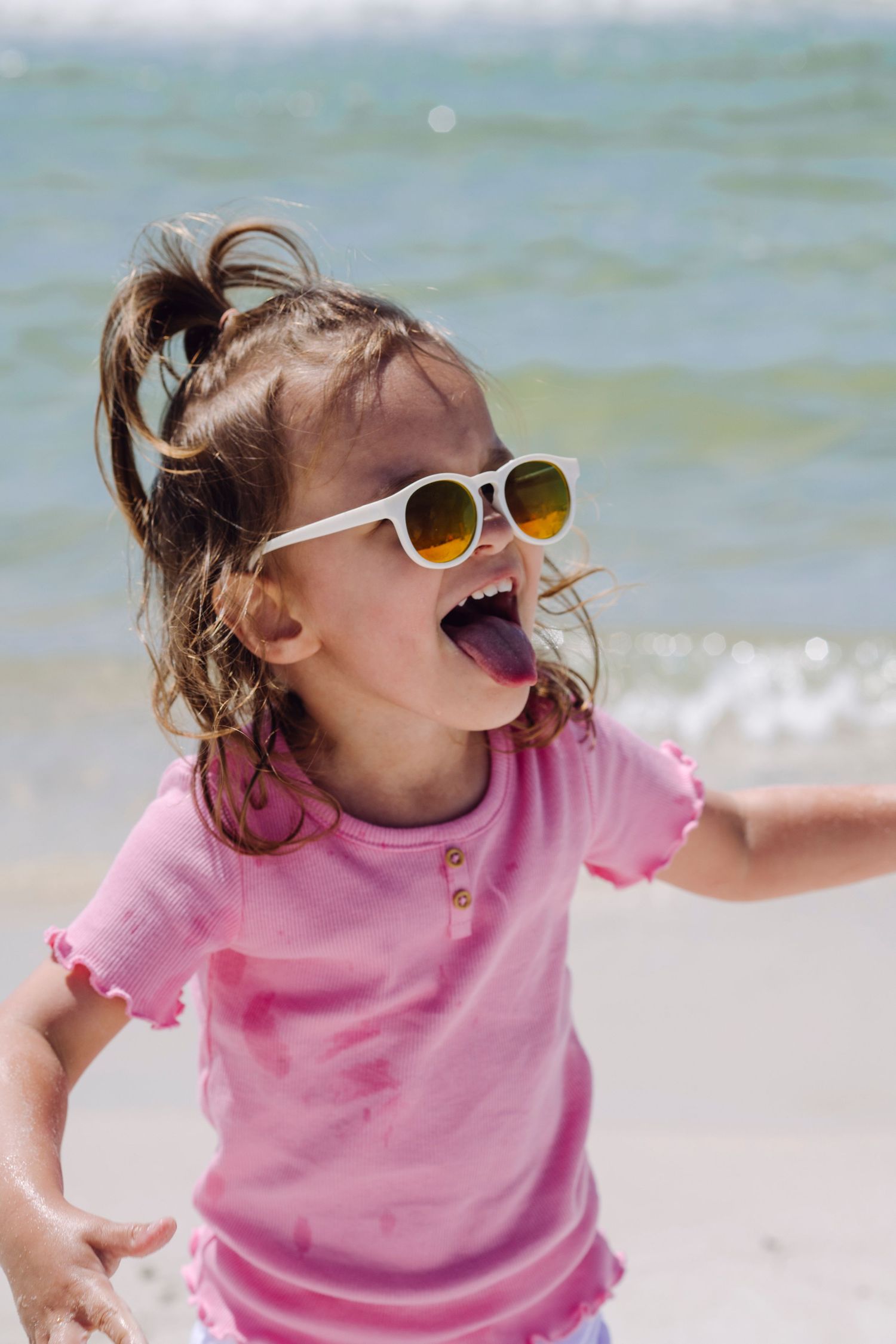Young child in pink shirt and white sunglasses laughs joyfully while playing at the beach on a sunny day.