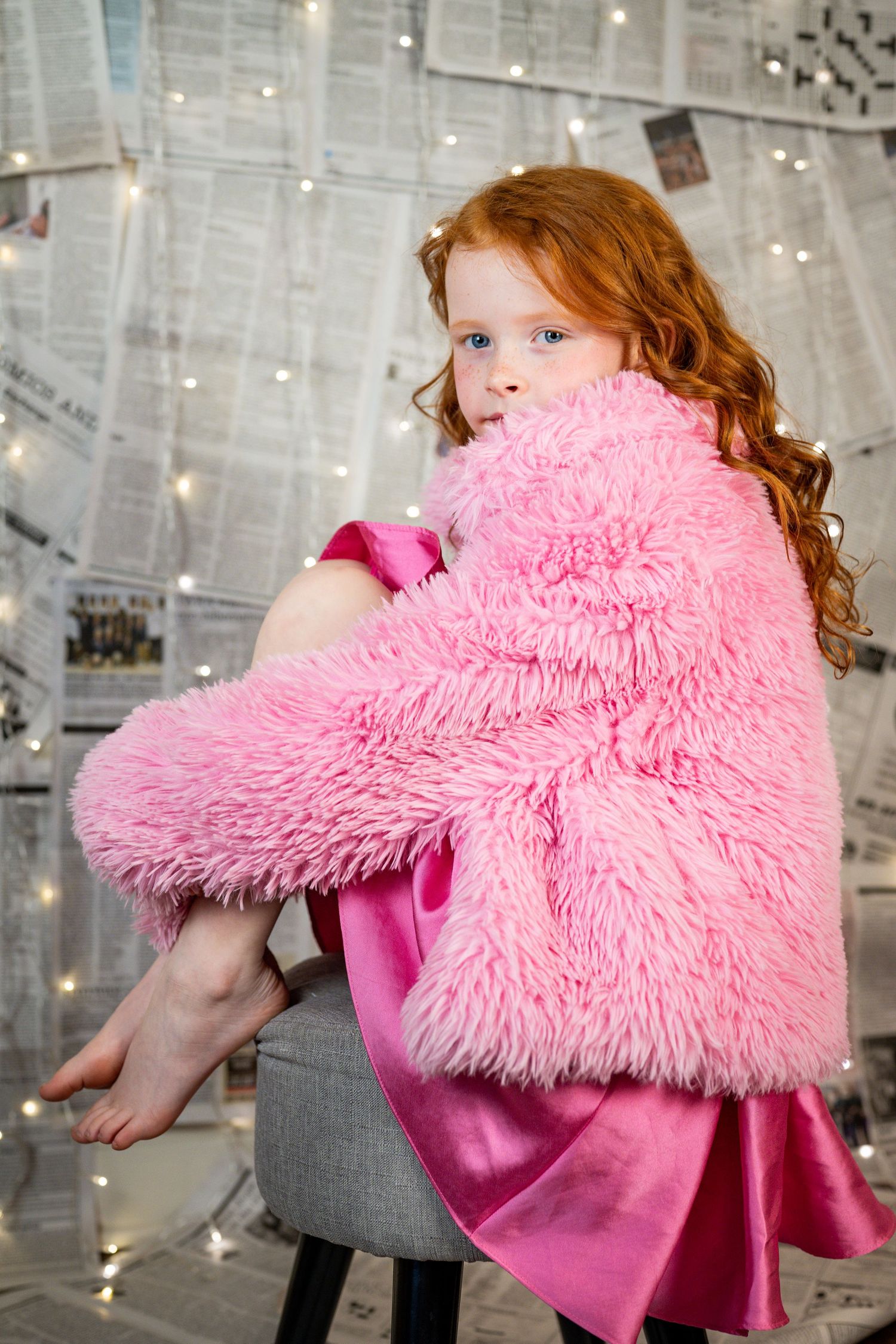 Redhead girl in pink fluffy coat and dress sits on stool against newspaper wall with fairy lights.