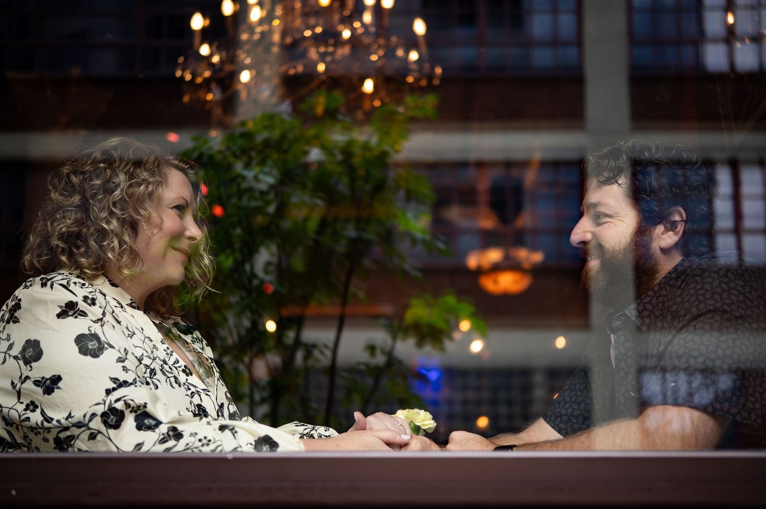 Couple sitting across from each other at a restaurant table, viewed through a window with warm ambient lighting.