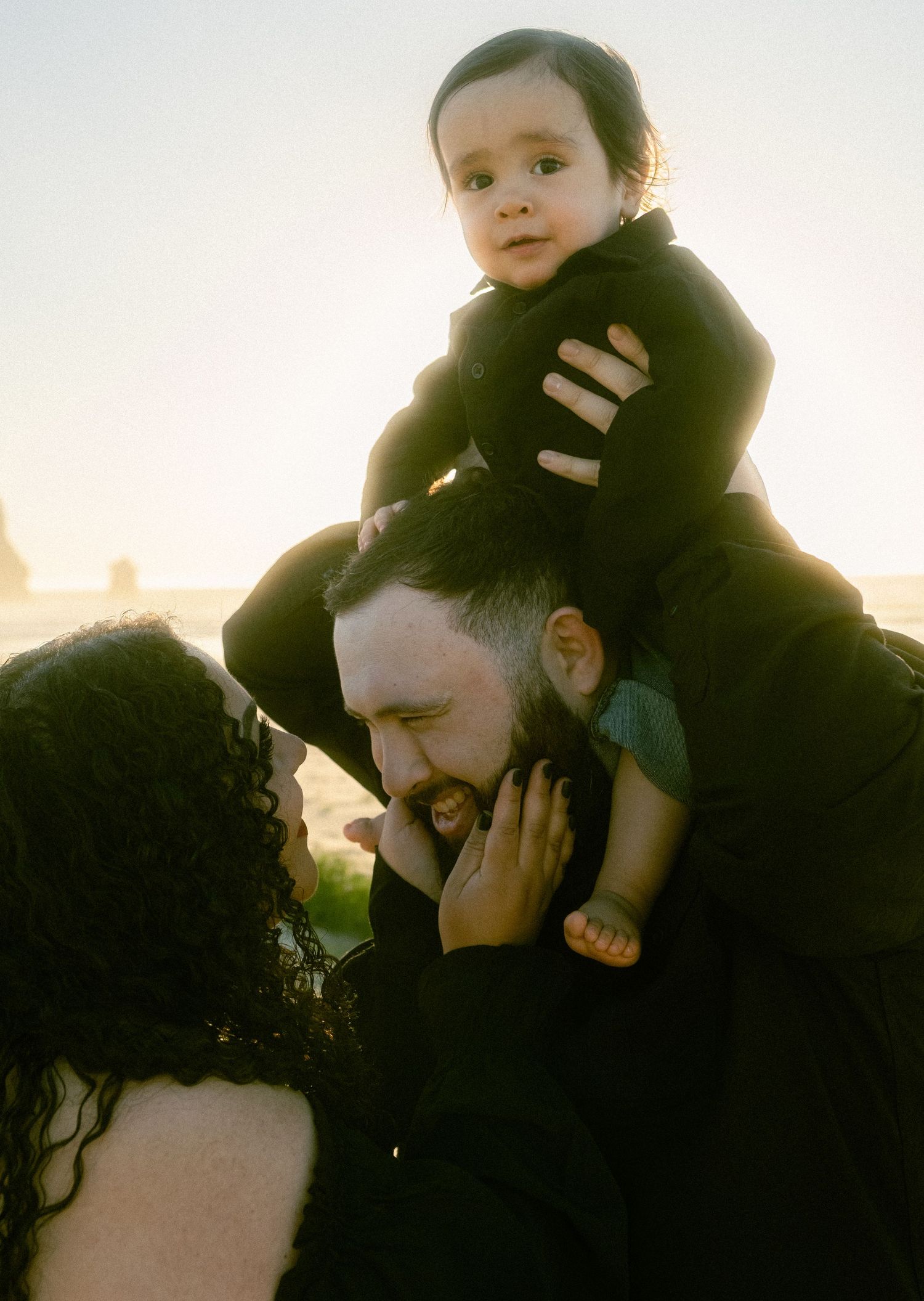 A family in black clothing poses at sunset on a beach, with a father lifting a baby above his head while a mother looks on.