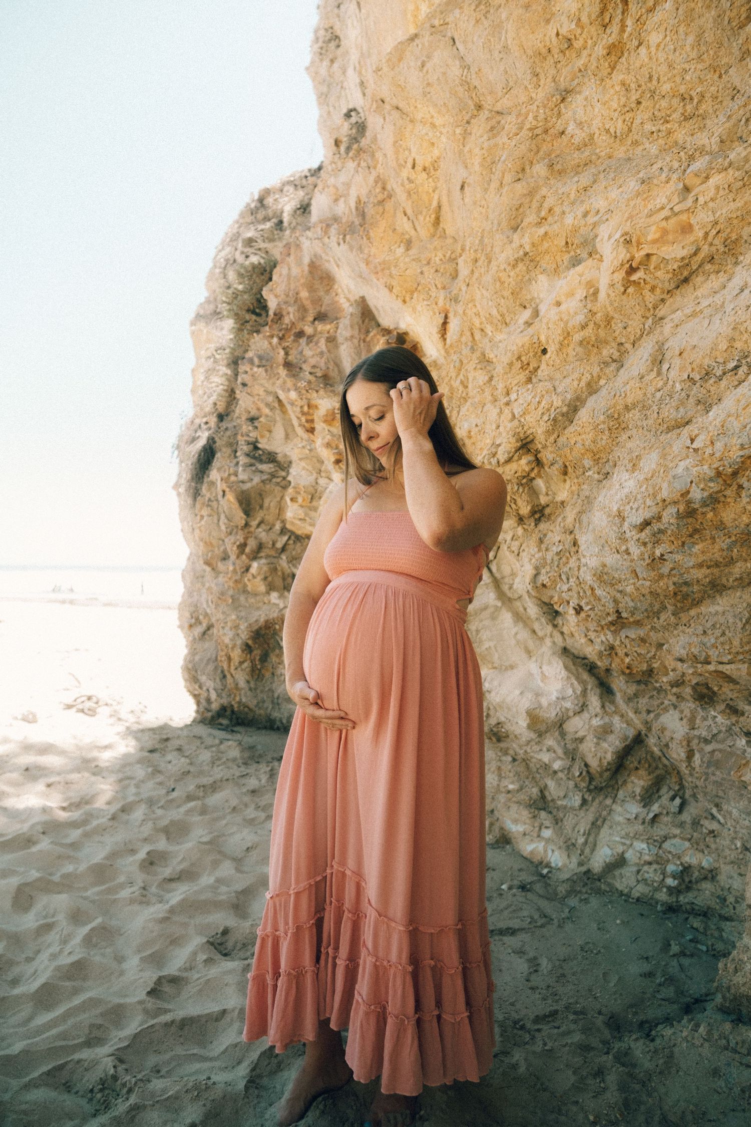 Pregnant woman in pink maxi dress posing near rocky beach cliff, cradling baby bump in serene maternity photo.