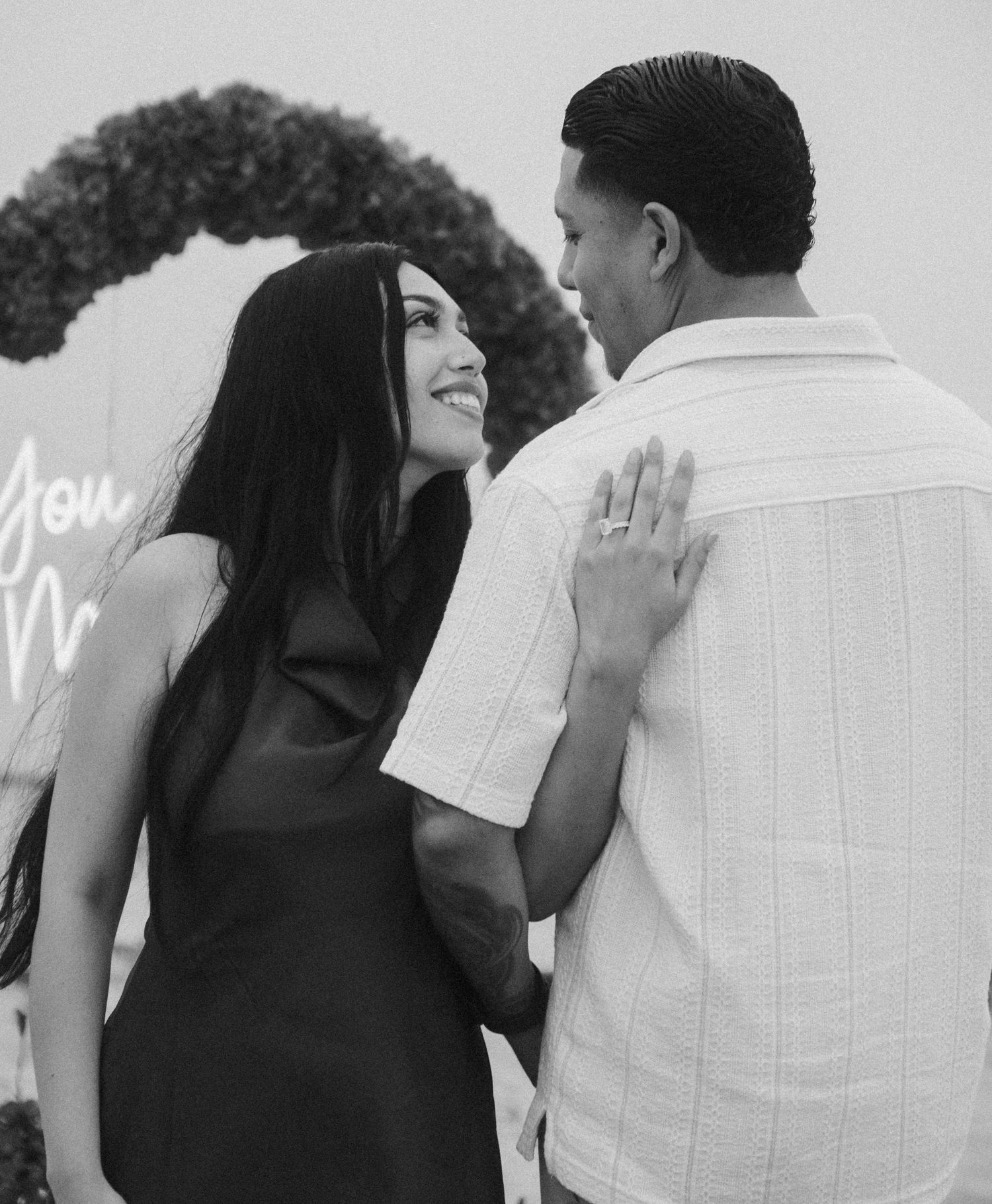 Black and white photo of a smiling couple at an engagement party, woman in black dress touching man's shoulder.