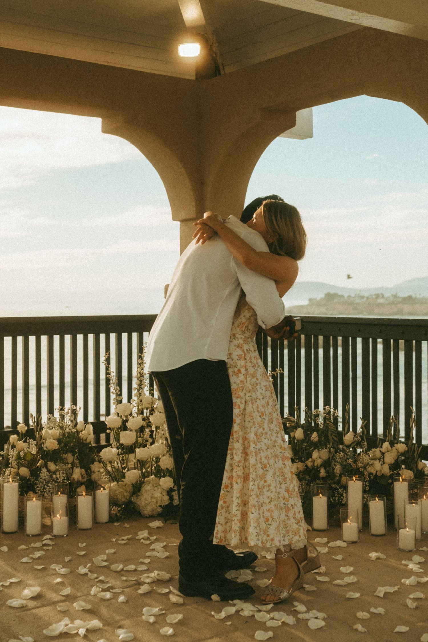 Couple embracing on a romantic candlelit balcony with floral arrangements and ocean views during a proposal.