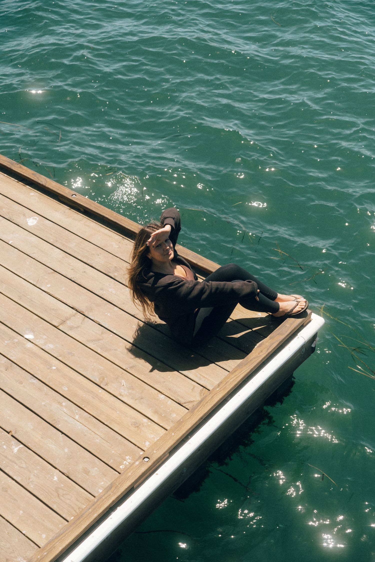 Woman in black outfit sitting on wooden dock edge over sparkling turquoise water on a sunny day.