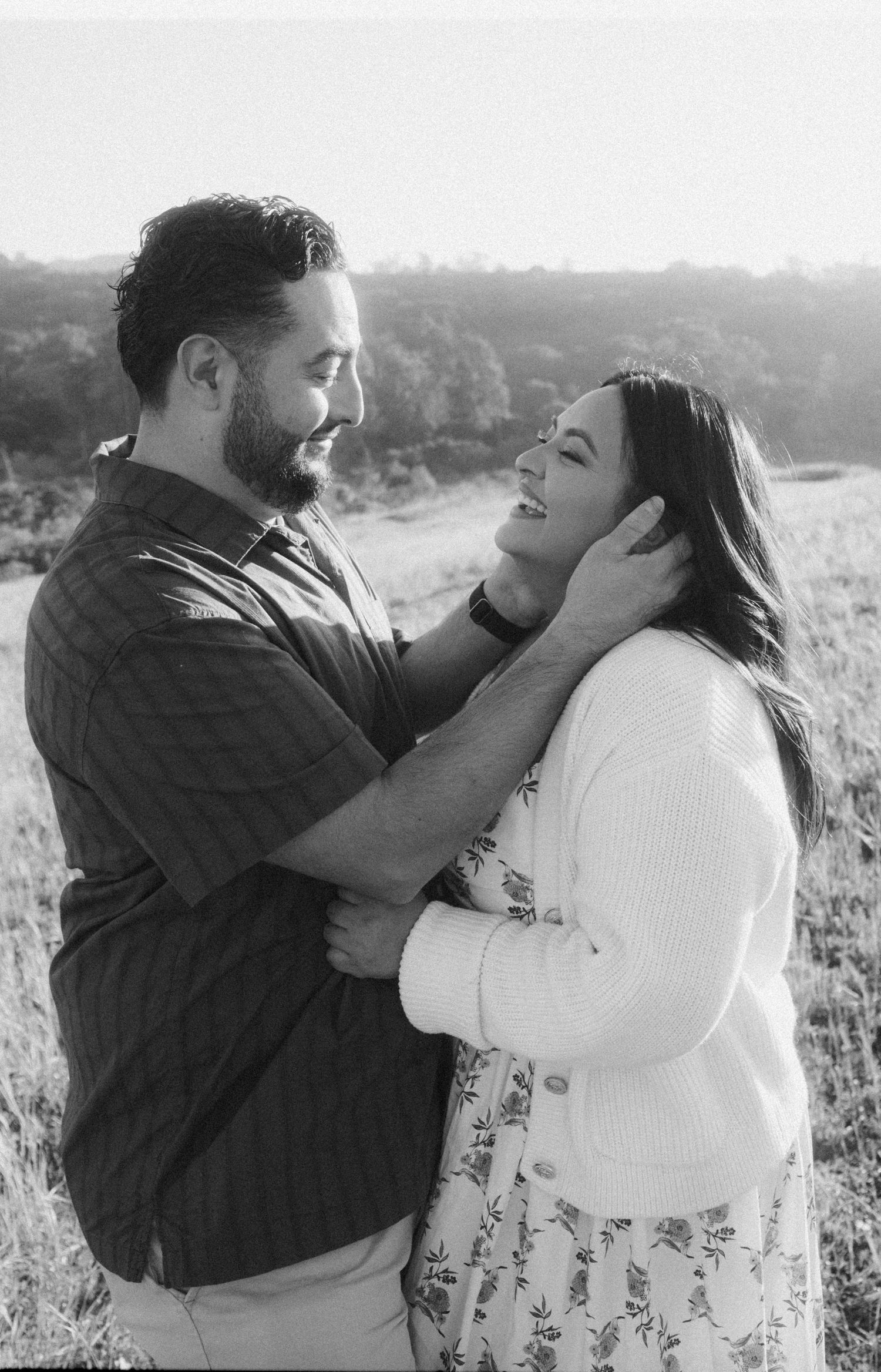 Black and white photo of a loving couple embracing in a field, smiling at each other during a romantic outdoor engagement session.