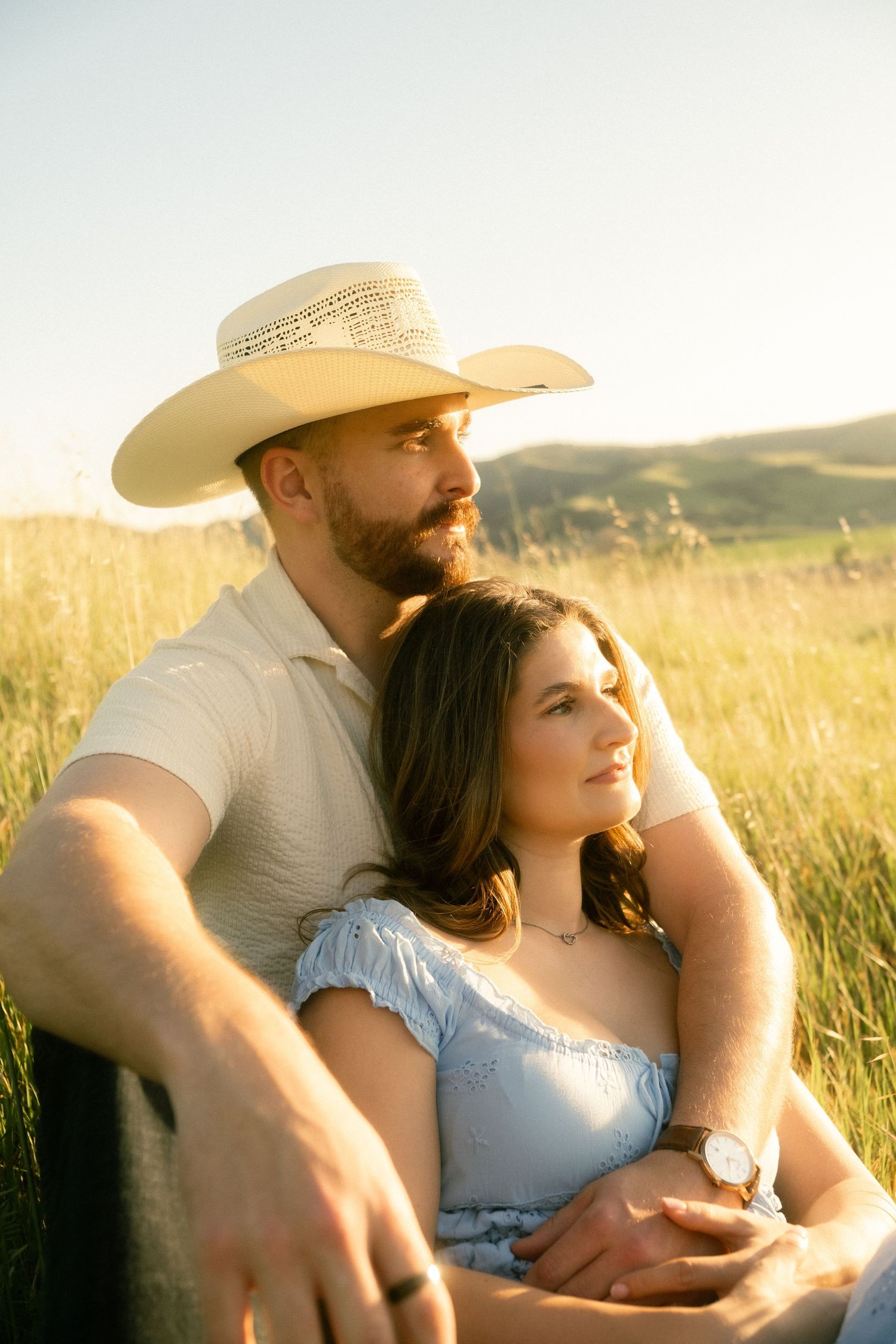 A couple sits together in a golden sunlit field, man in cowboy hat embracing woman in blue dress.