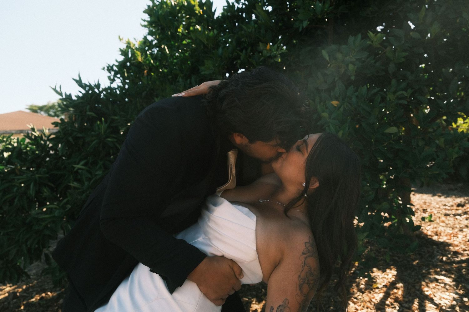 Wedding couple poses against lush green trees in outdoor setting.