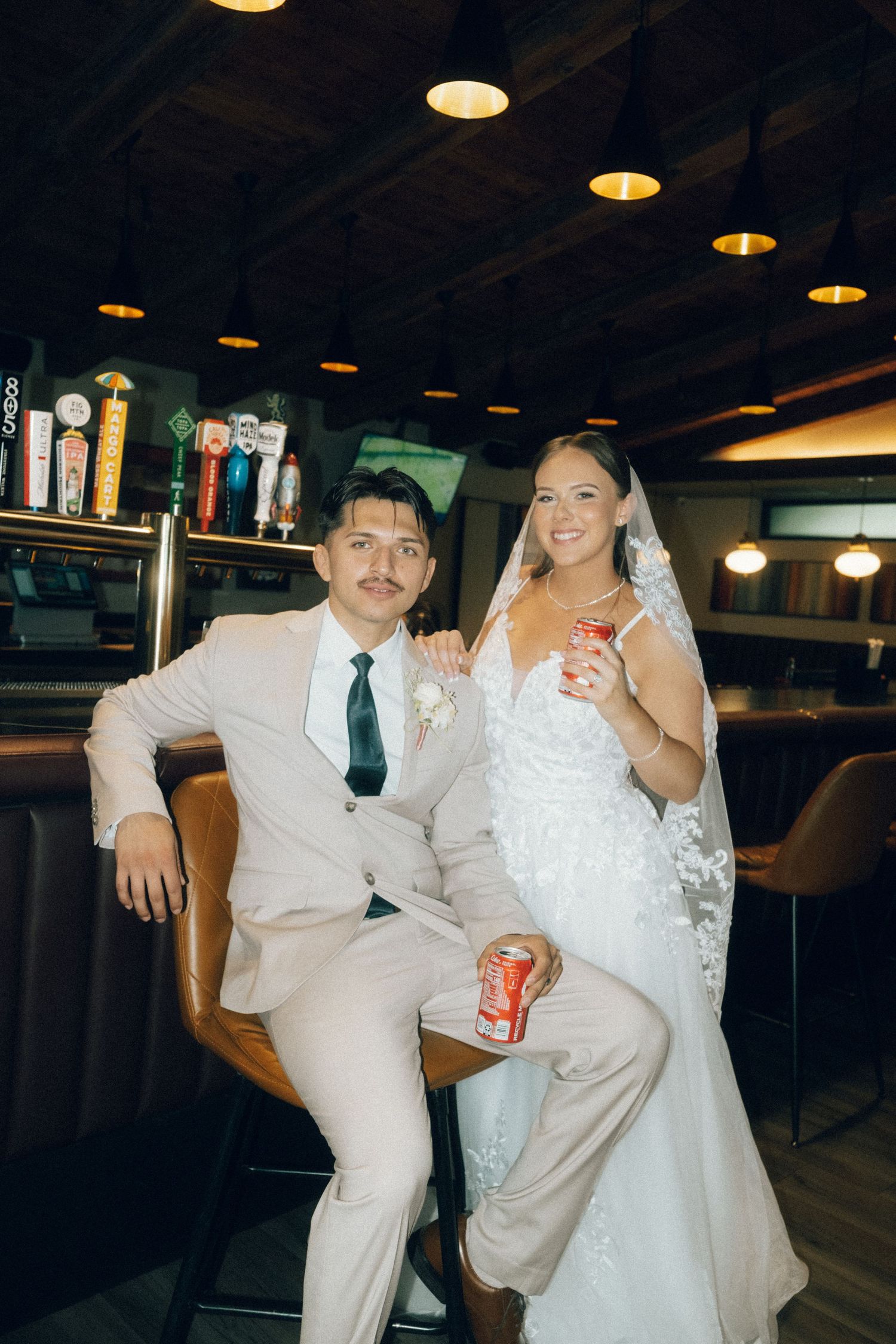 Bride and groom posing together at a bar, holding Coca-Cola cans, dressed in wedding attire with veil and suit.