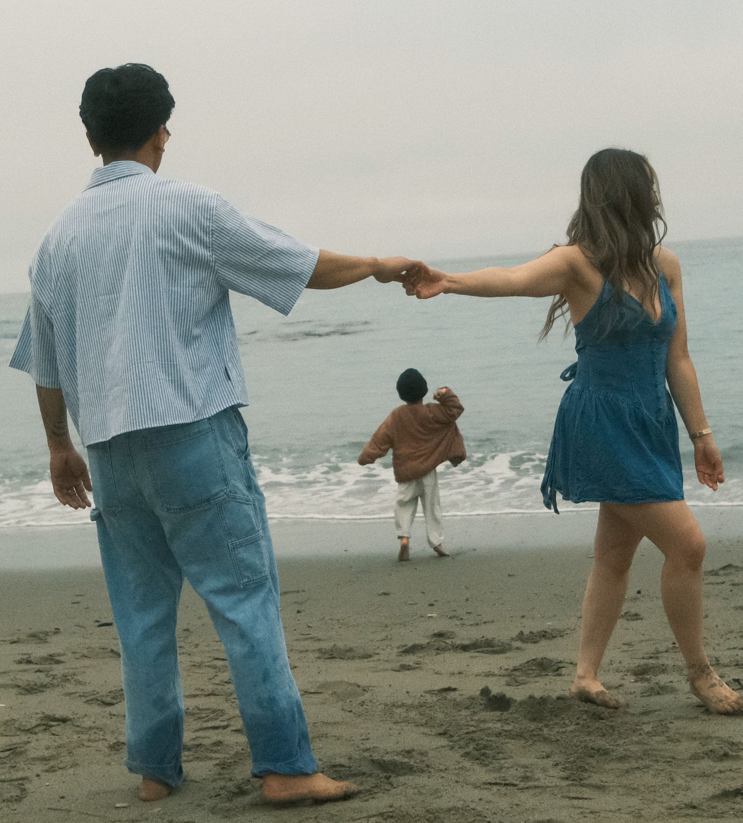 A couple holds hands on a misty beach while a child runs toward the ocean waves.