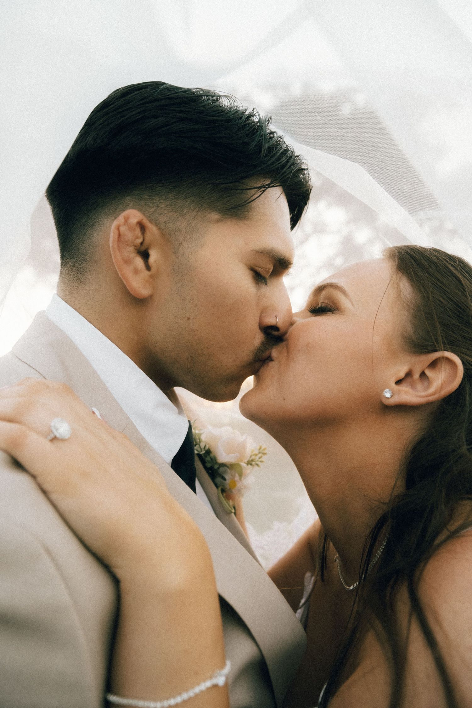 Bride and groom sharing a romantic kiss under a flowing veil on their wedding day.