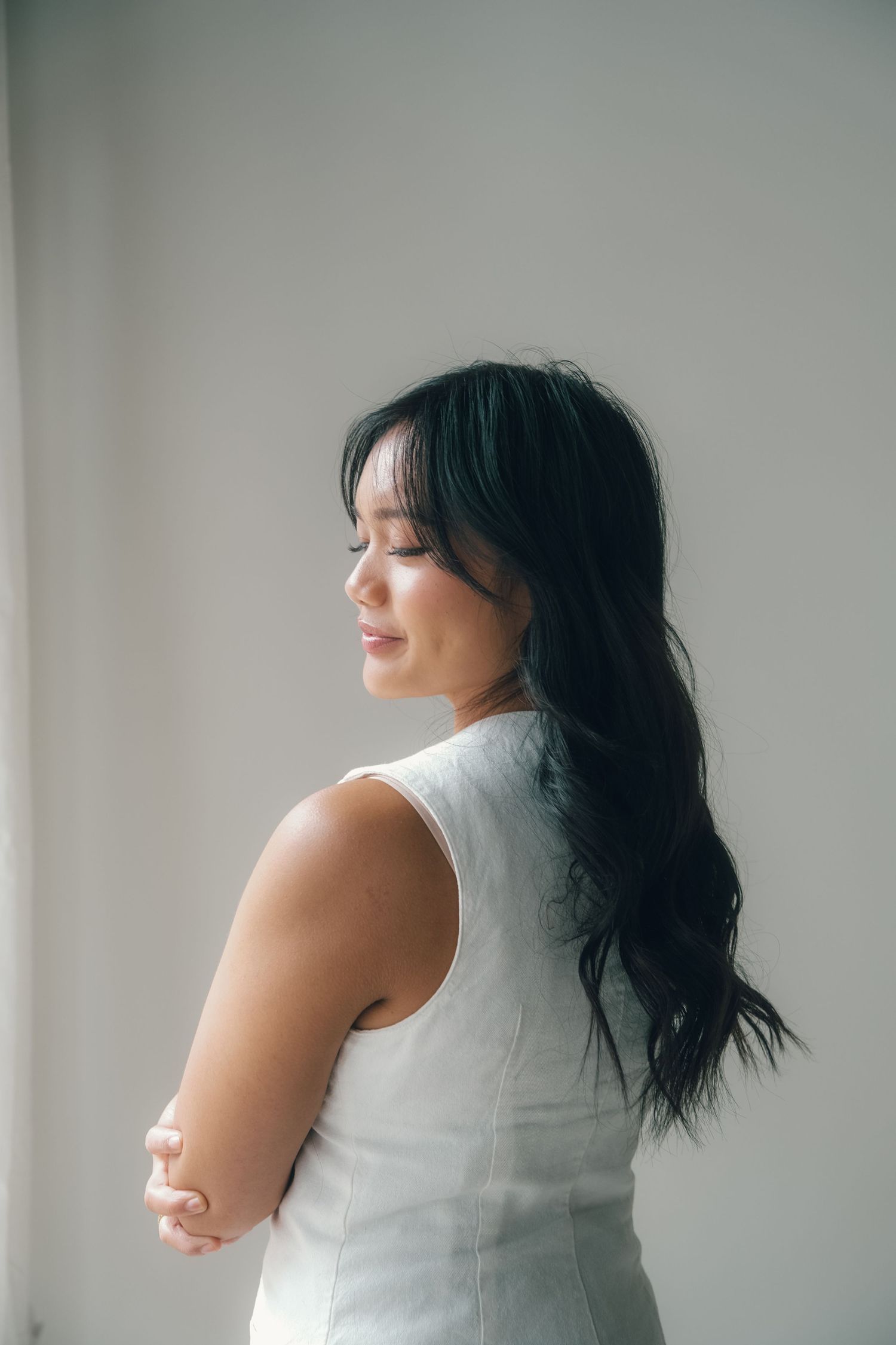 A young woman with long dark hair wearing a white sleeveless dress, smiling softly with eyes closed against a light background.