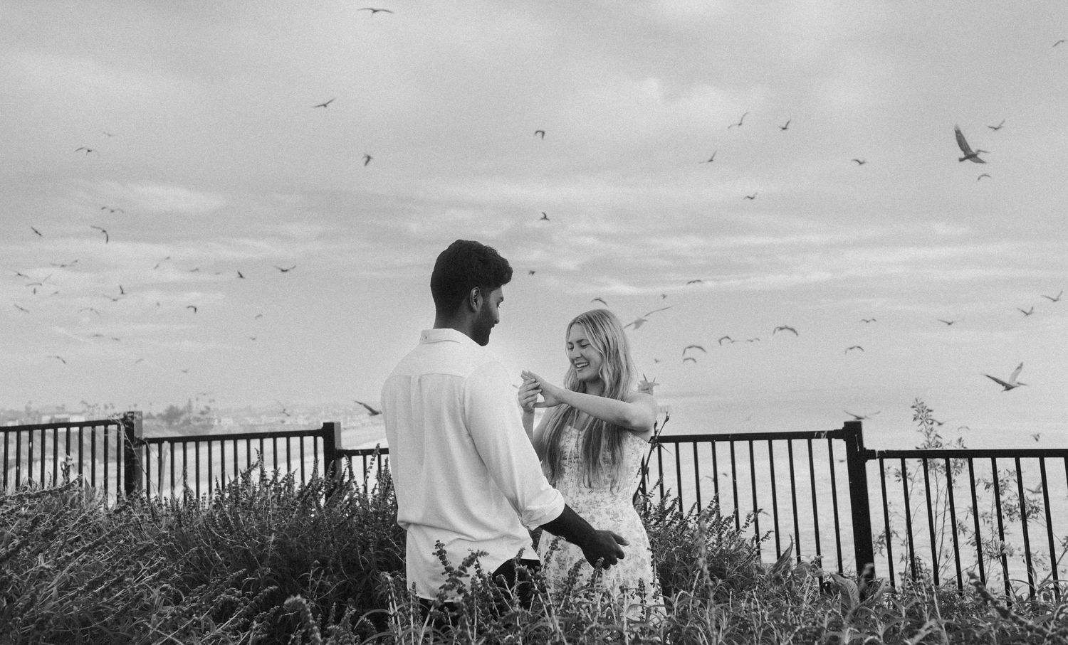 Couple dancing romantically outdoors near a fence with birds flying in a black and white photo.
