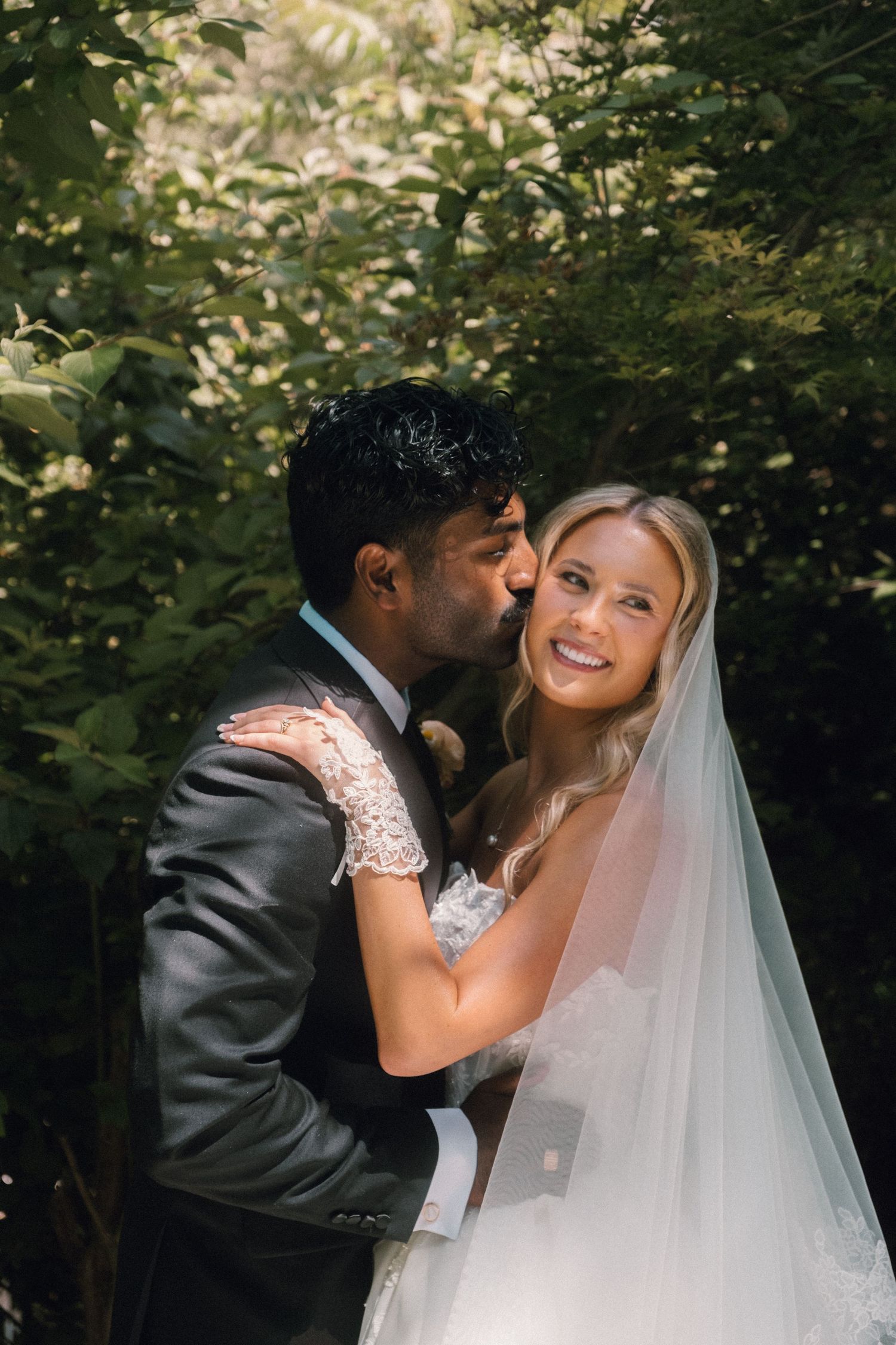 Groom kissing bride on cheek outdoors, bride smiling in white gown and veil, surrounded by lush green foliage.