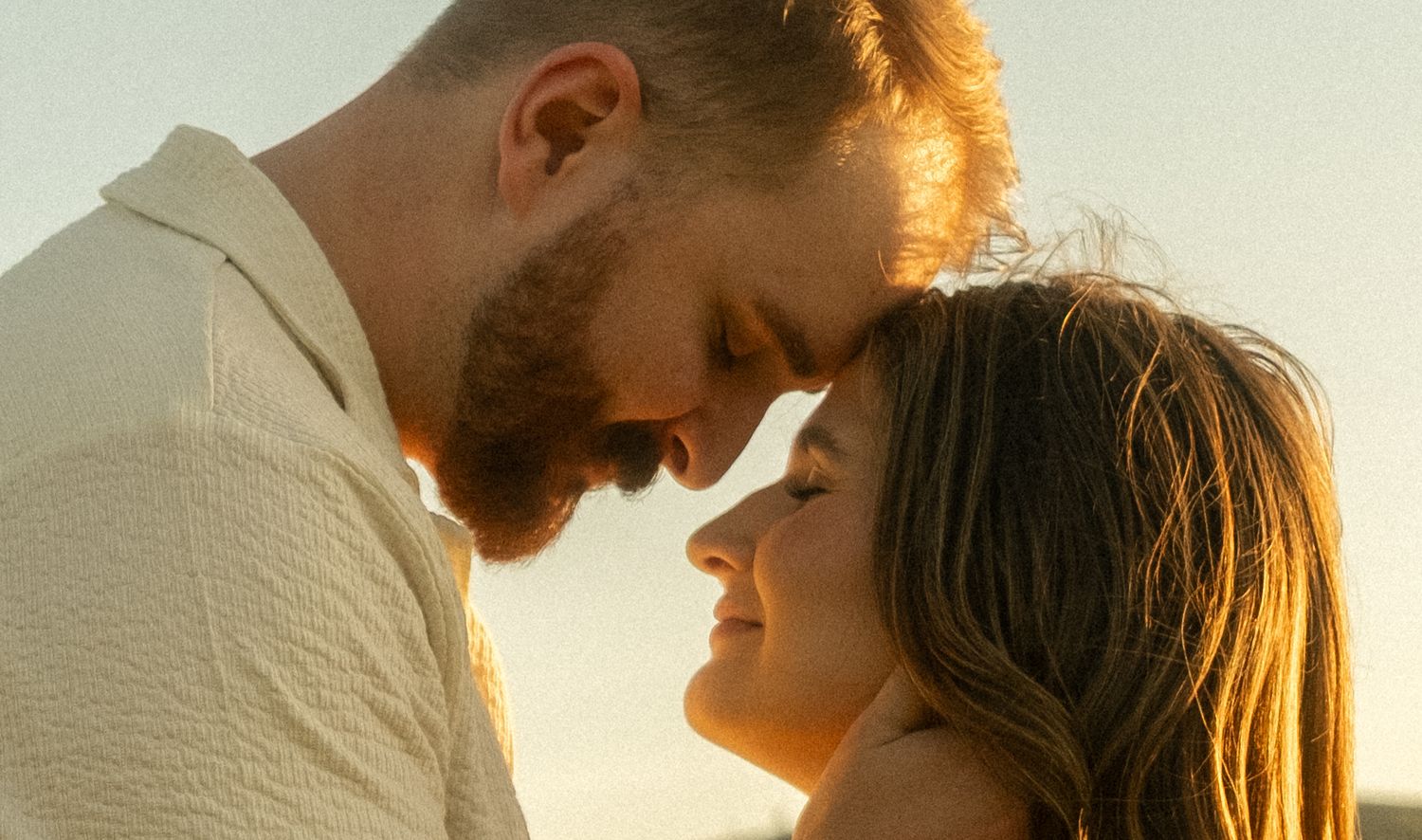 Romantic couple sharing an intimate moment at golden hour, foreheads nearly touching in warm sunset light.