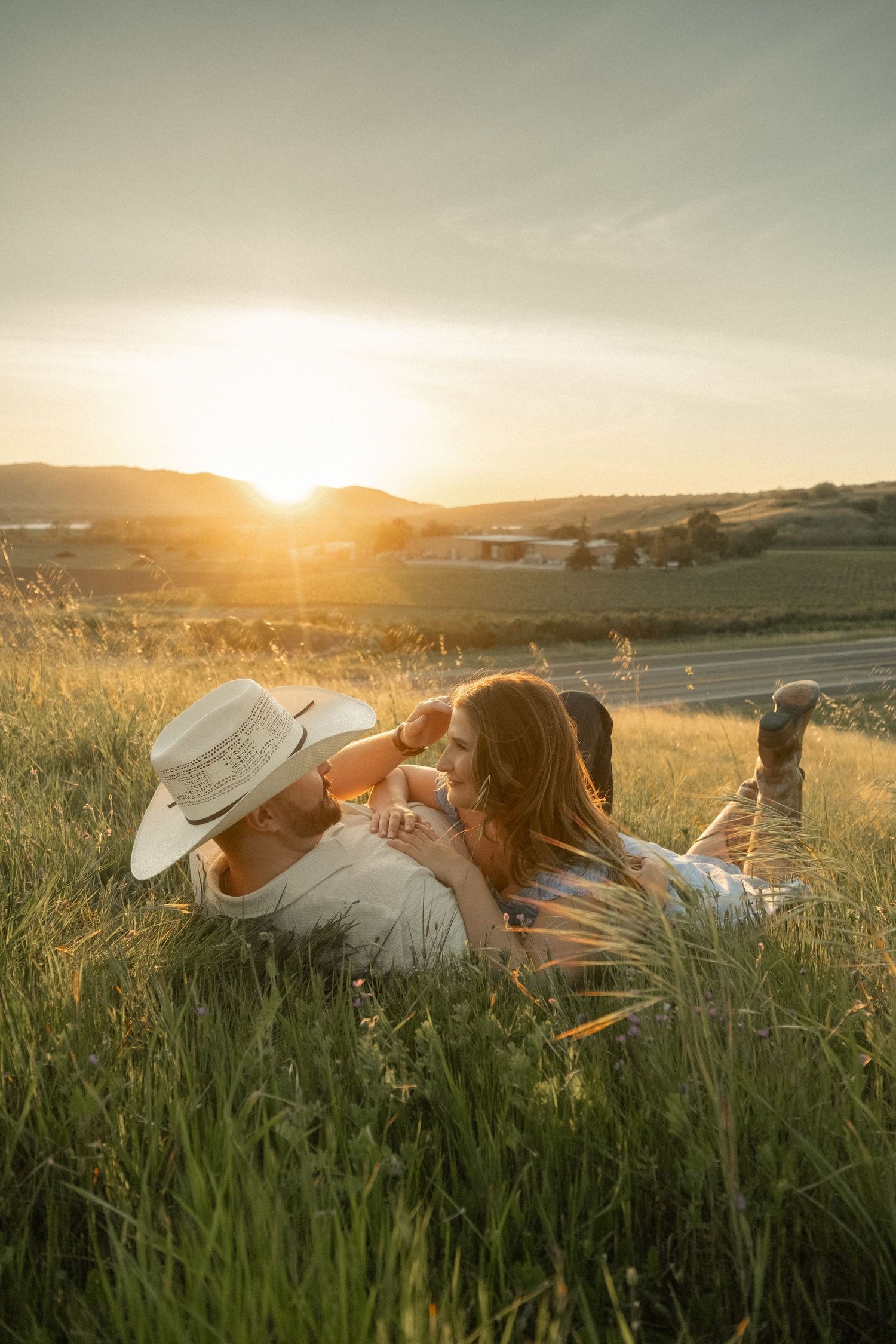 Couple lying in golden grass field at sunset, woman resting on man wearing cowboy hat, rolling hills backdrop.
