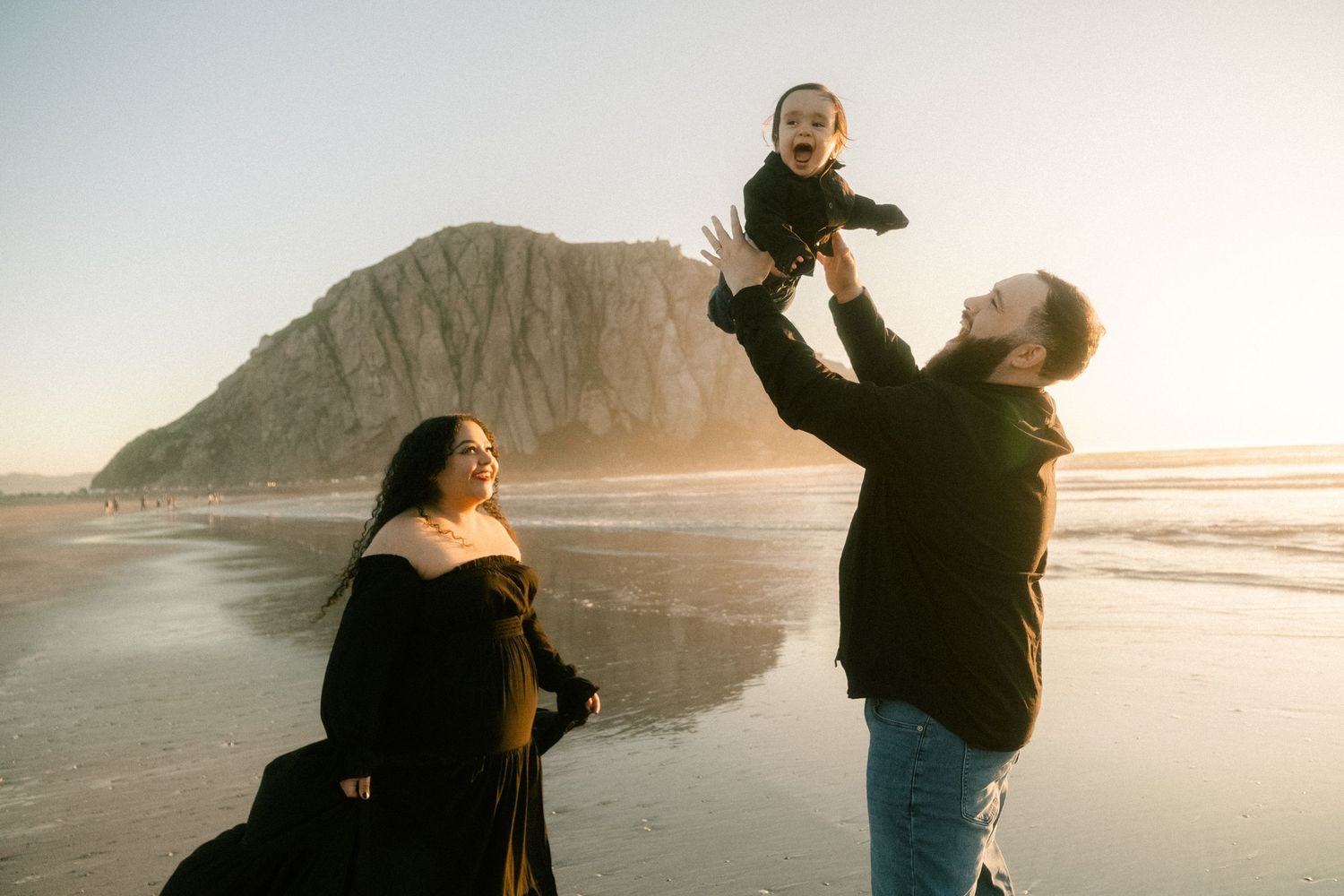 Family enjoying a beach day at Morro Rock, parents lifting laughing baby in the air at golden hour.