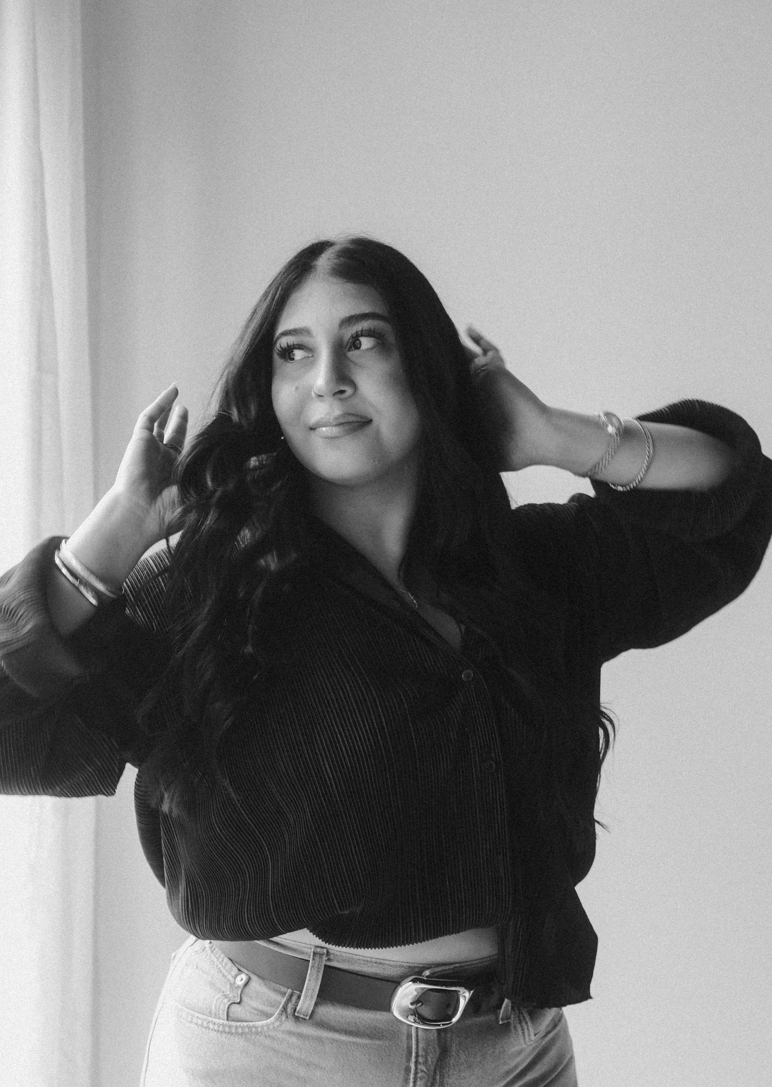 Black and white photo of a young woman with long wavy hair posing with hands in hair, wearing a dark top and jeans.