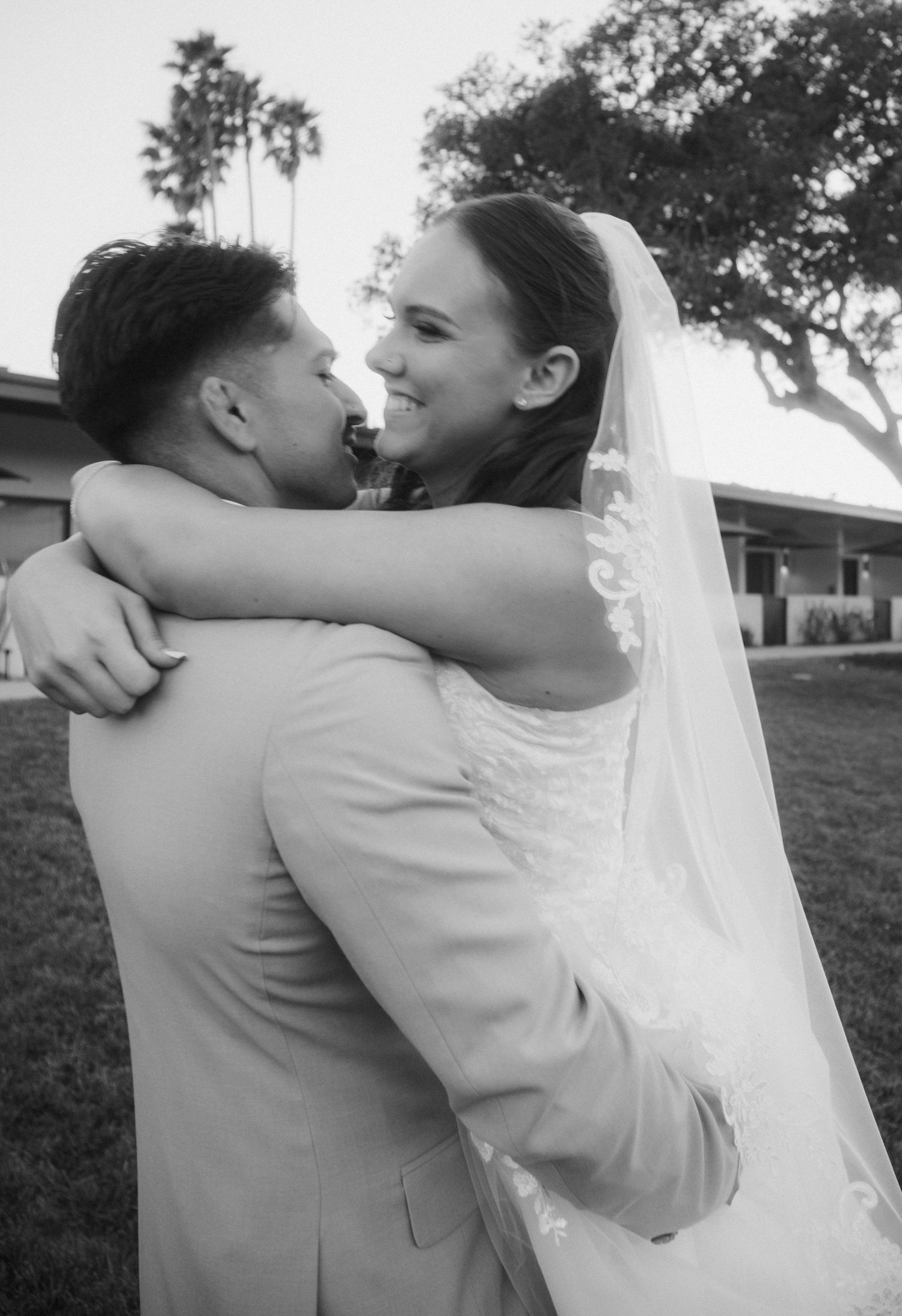 Black and white photo of a smiling bride in veil embracing her groom outdoors on their wedding day.